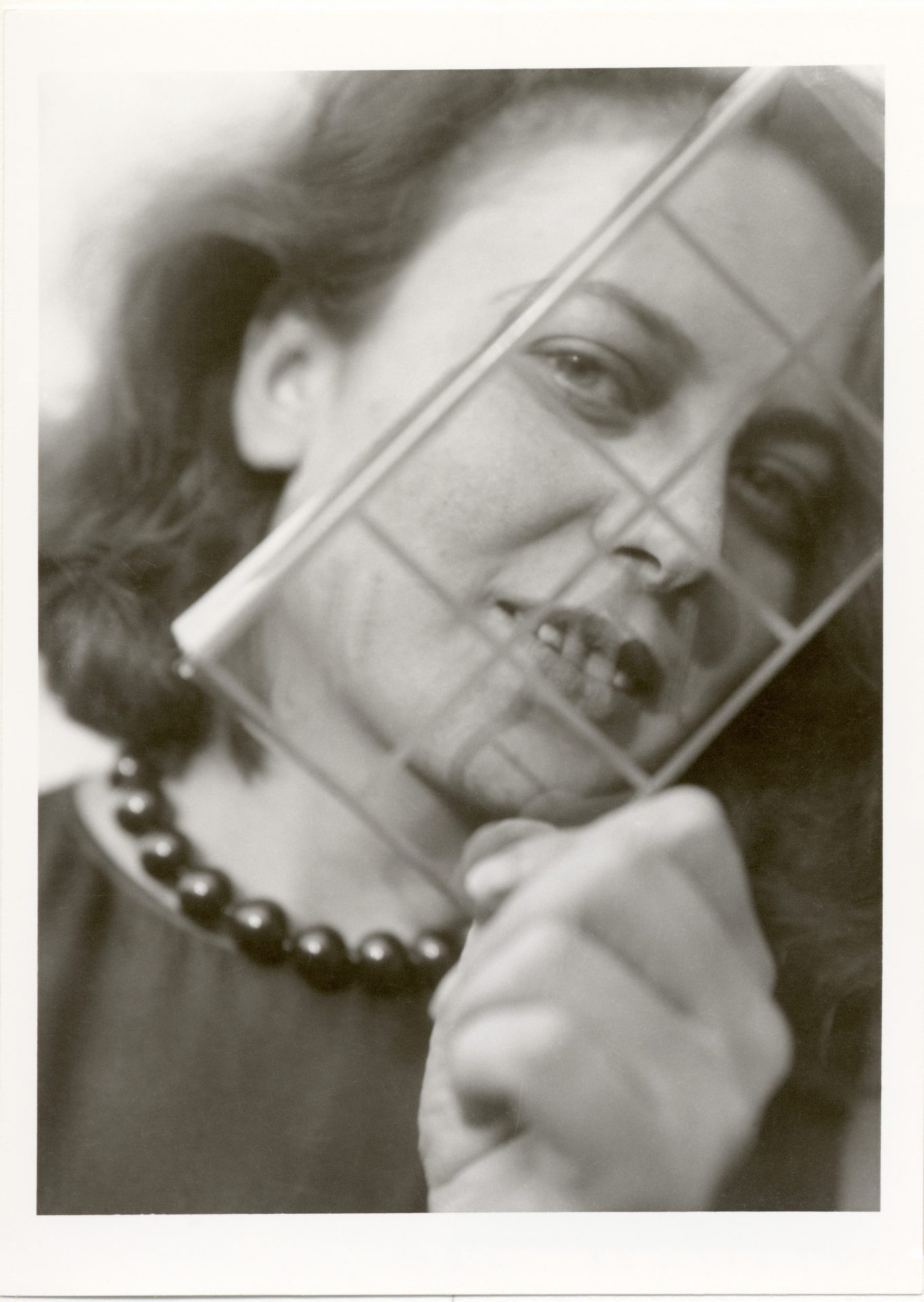 Schwarz-weiß-Foto einer lächelnden Frau mit einer dunklen Perlenkette, die einen durchsichtigen Gitterrahmen vor ihr Gesicht hält.