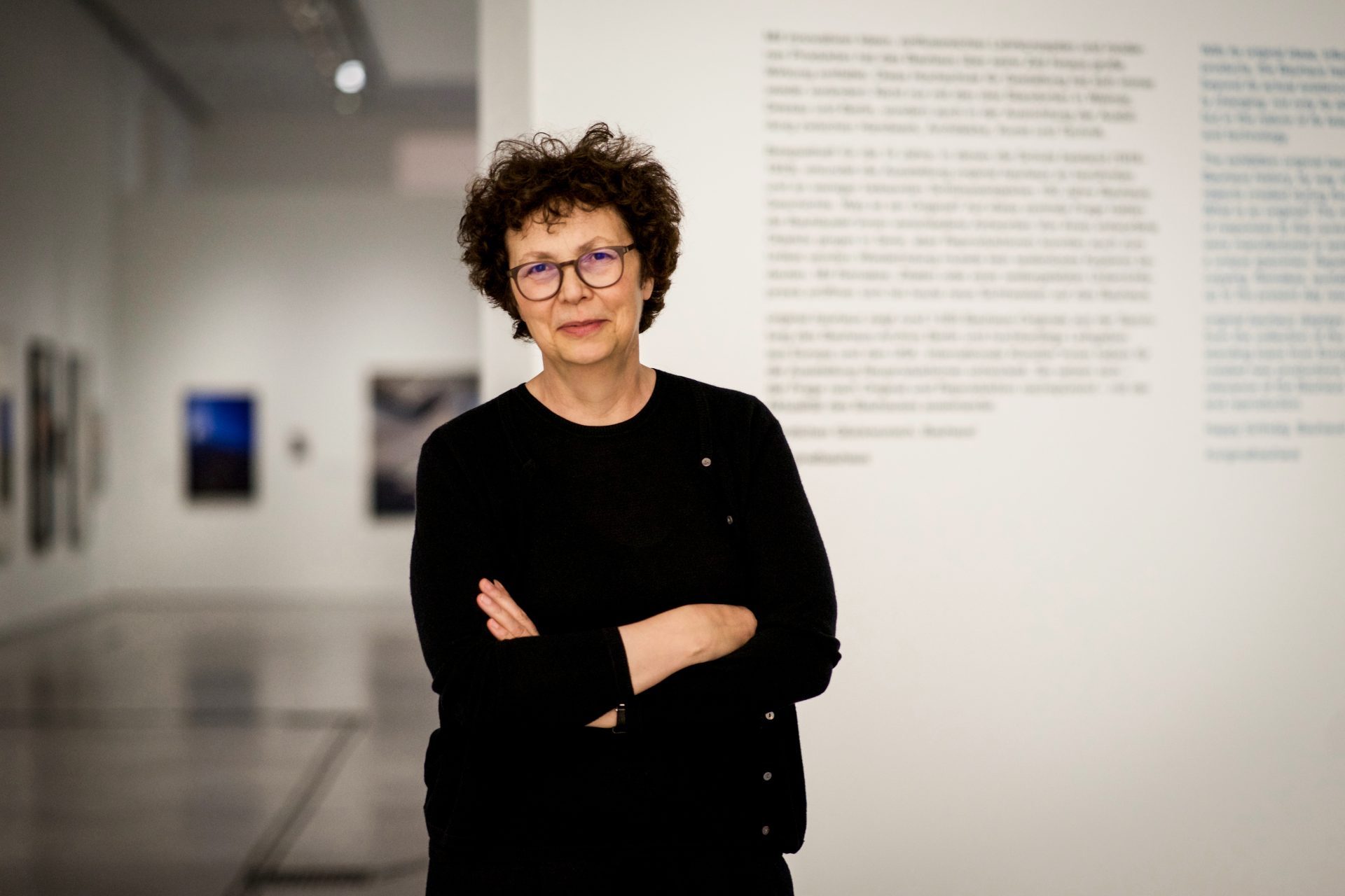 Foto von Dr. Annemarie Jaeggi, wie sie lächelnd und mit verschränkten Armen in einem hellen Ausstellungsraum vor Wandtext steht.