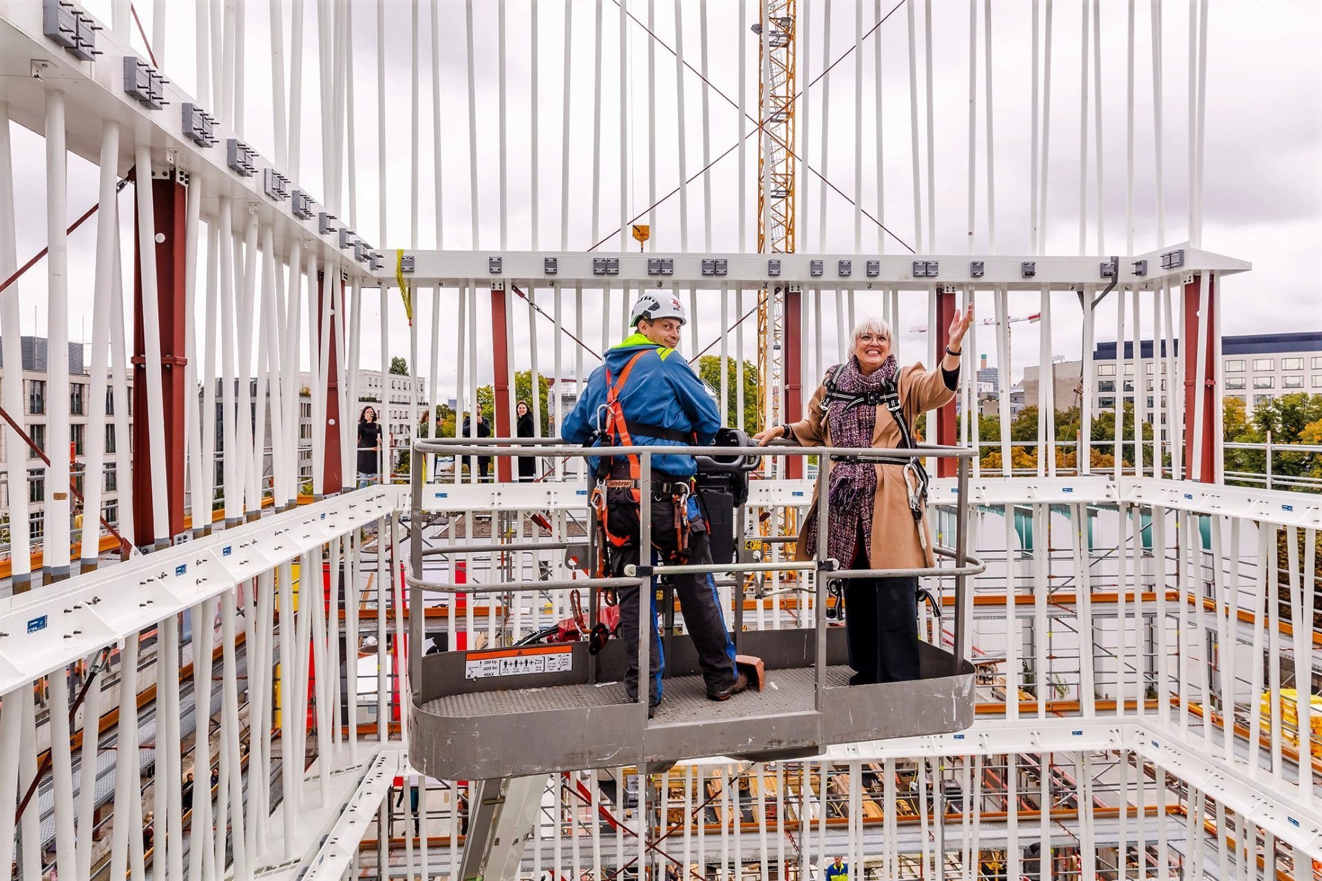 Zwei Personen stehen auf einem Gerüst auf der Baustelle, umgeben von weiteren Gerüsten und einem Kran im Hintergrund.