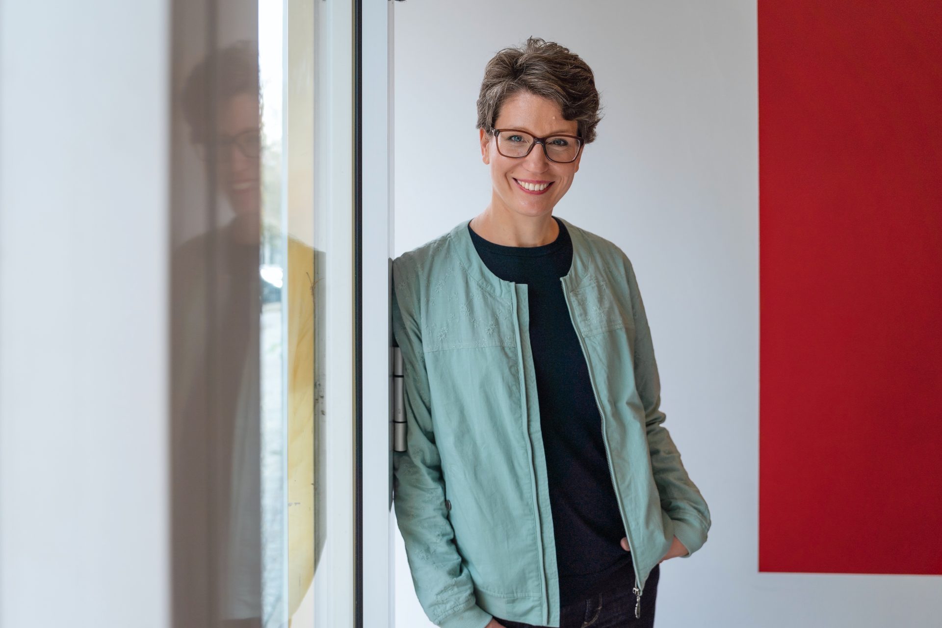 Foto einer lächelnden Person mit Brille, steht an einer Tür und trägt eine graue Jacke vor modernem roten Hintergrund.