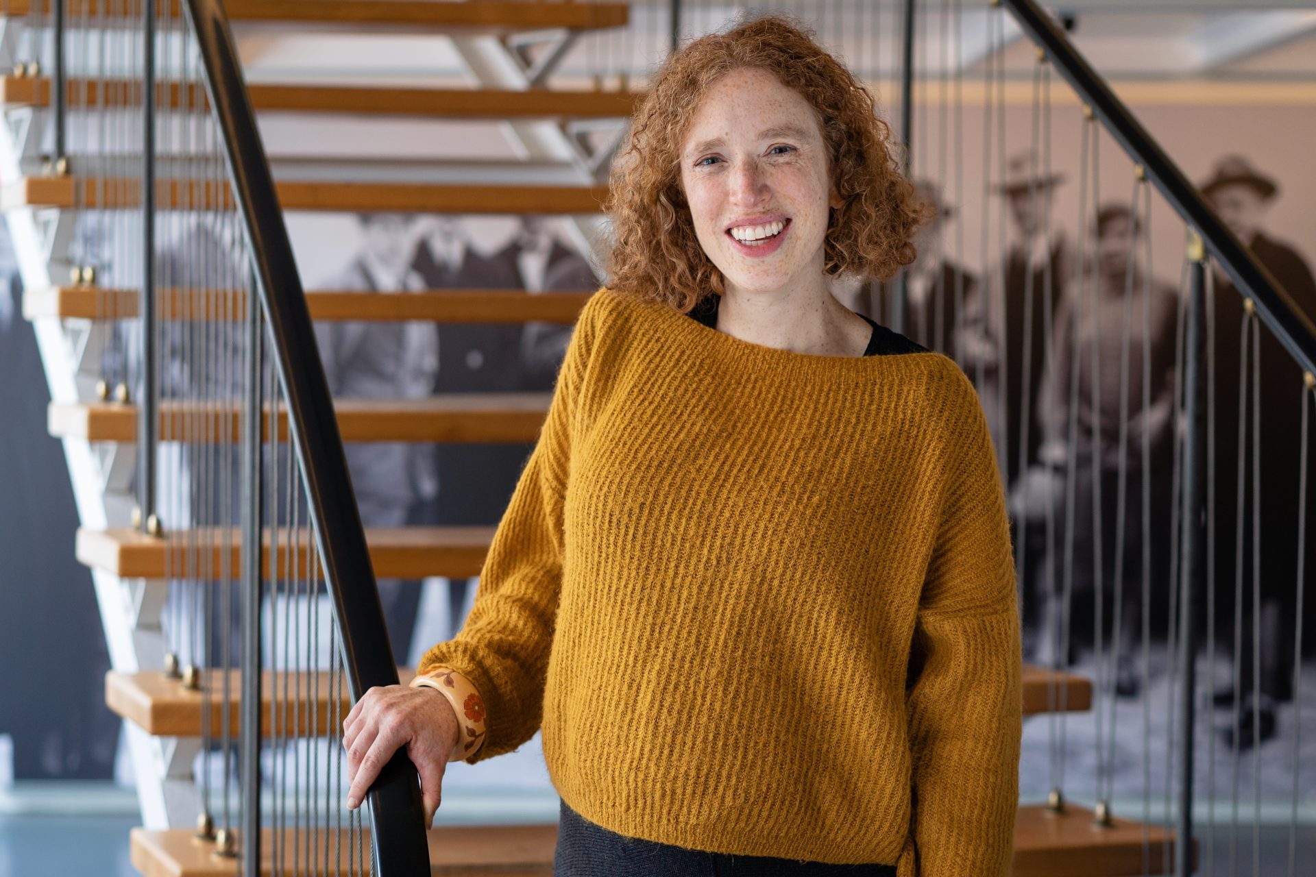 Foto einer Person mit lockigem, rotem Haar, die einen orangefarbenen Pullover trägt und lächelt auf einer Treppe steht.