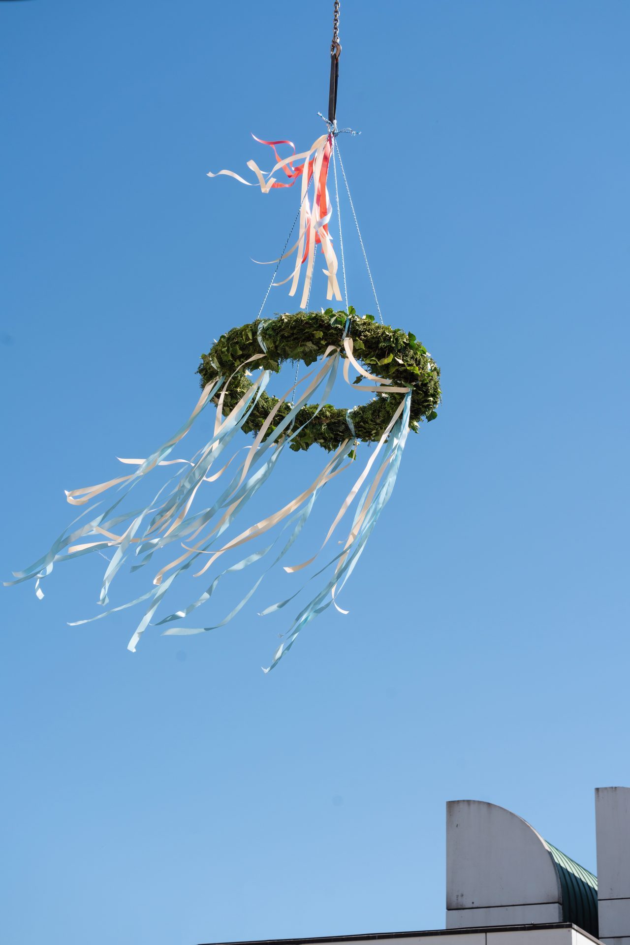 Grüner Kranz mit bunten Bändern schwebt vor blauem Himmel, an einem Seil hängend.