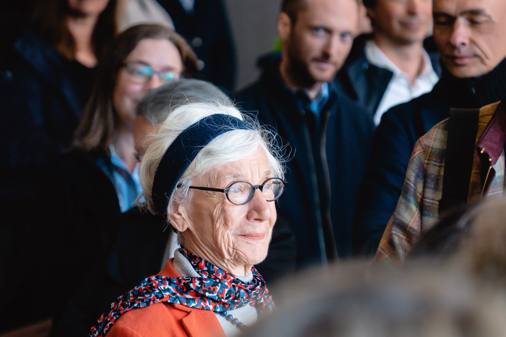 Ältere Frau mit Brille und grauen Haaren, umgeben von Menschen, wirkt nachdenklich und zufrieden.