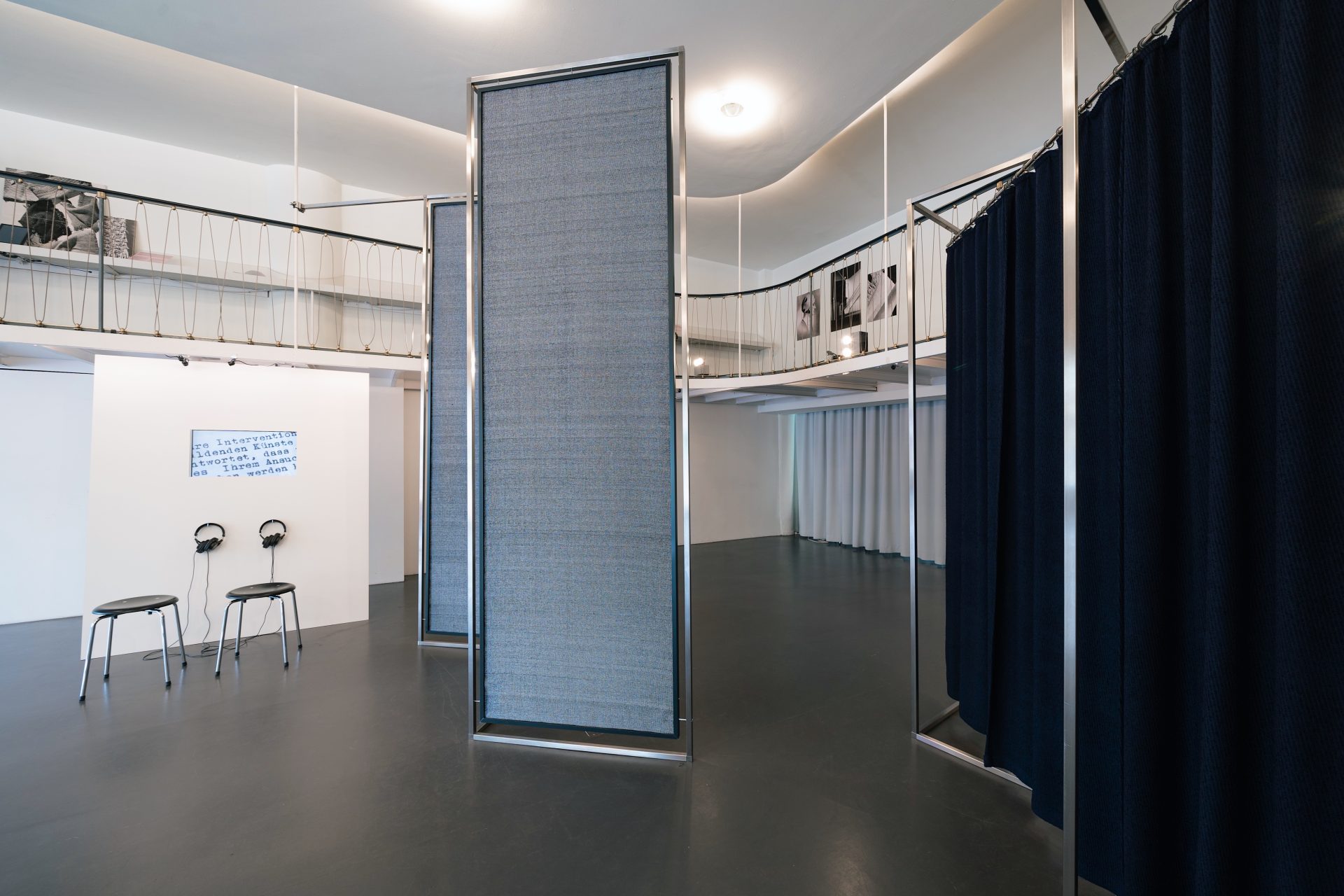 Foto des Innenraums im Temporary Bauhaus-Archiv: Graue und schwarze hohe Trennwände aus nachgewebten Stoffen, zwei schwarze Hocker vor einem Bildschrim und Blick auf die Galerieetage im Hintergrund.
