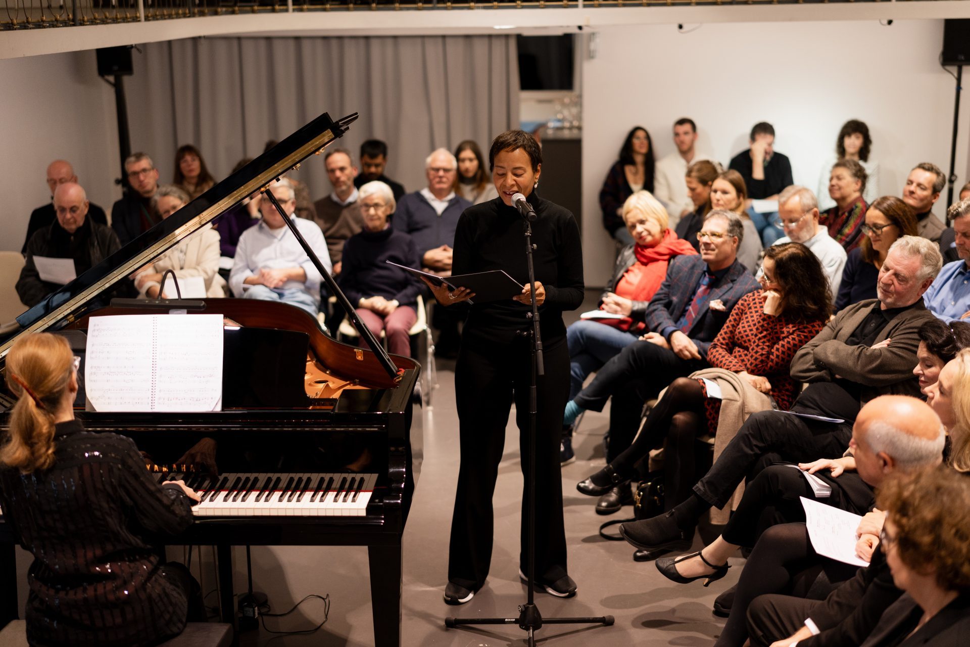 A piano is being played while a person reads or sings in front of an audience. The spectators are attentive and interested.
