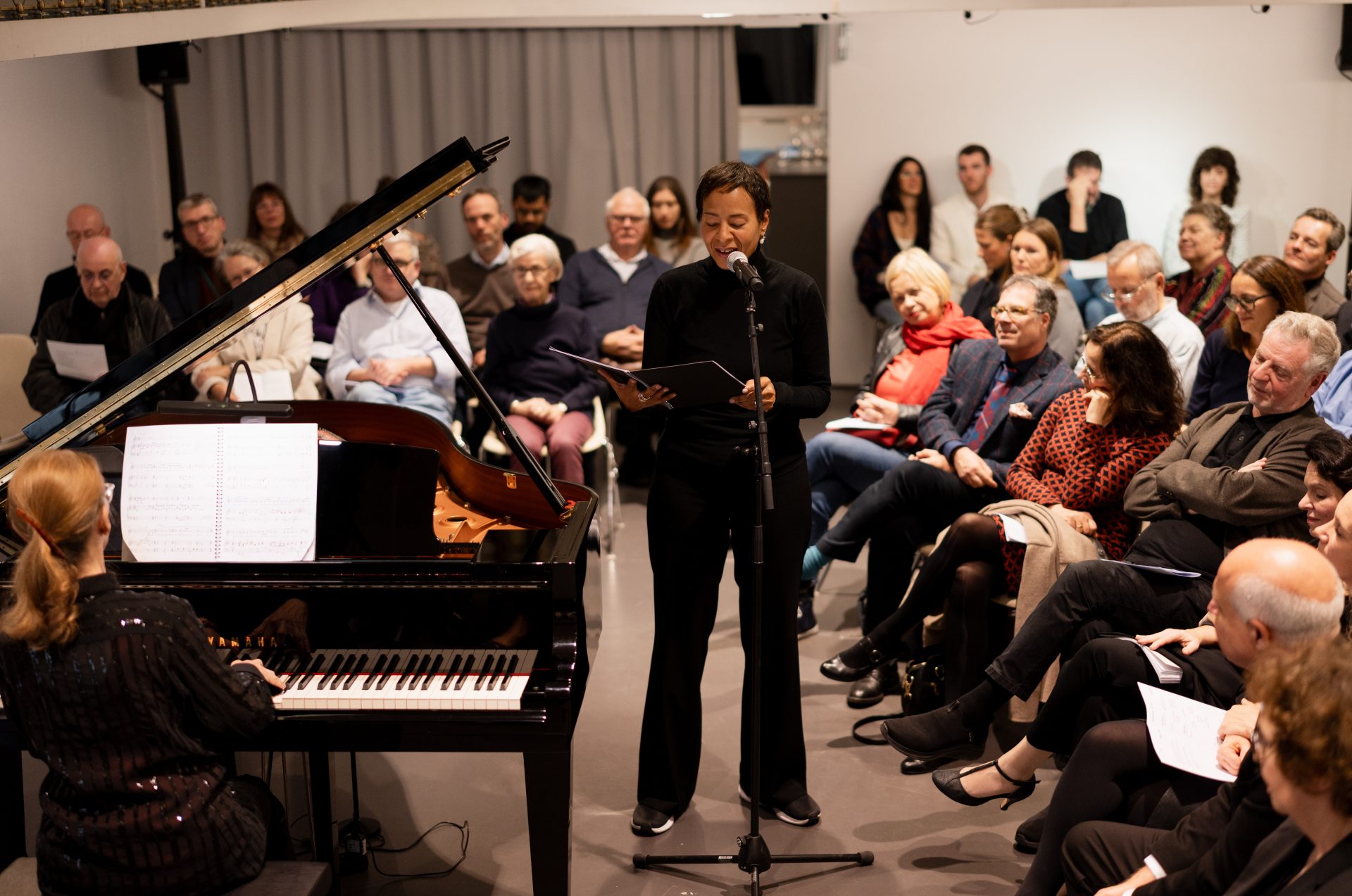 Ein Klavier wird gespielt, während jemand vor Publikum liest oder singt. Die Zuschauer sind aufmerksam und interessiert.