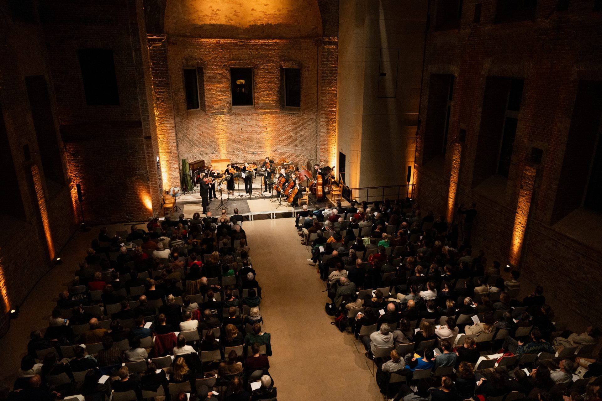 Konzert in einem alten, gut gefüllten Saal mit einer Band auf der Bühne und Publikum im Vordergrund.