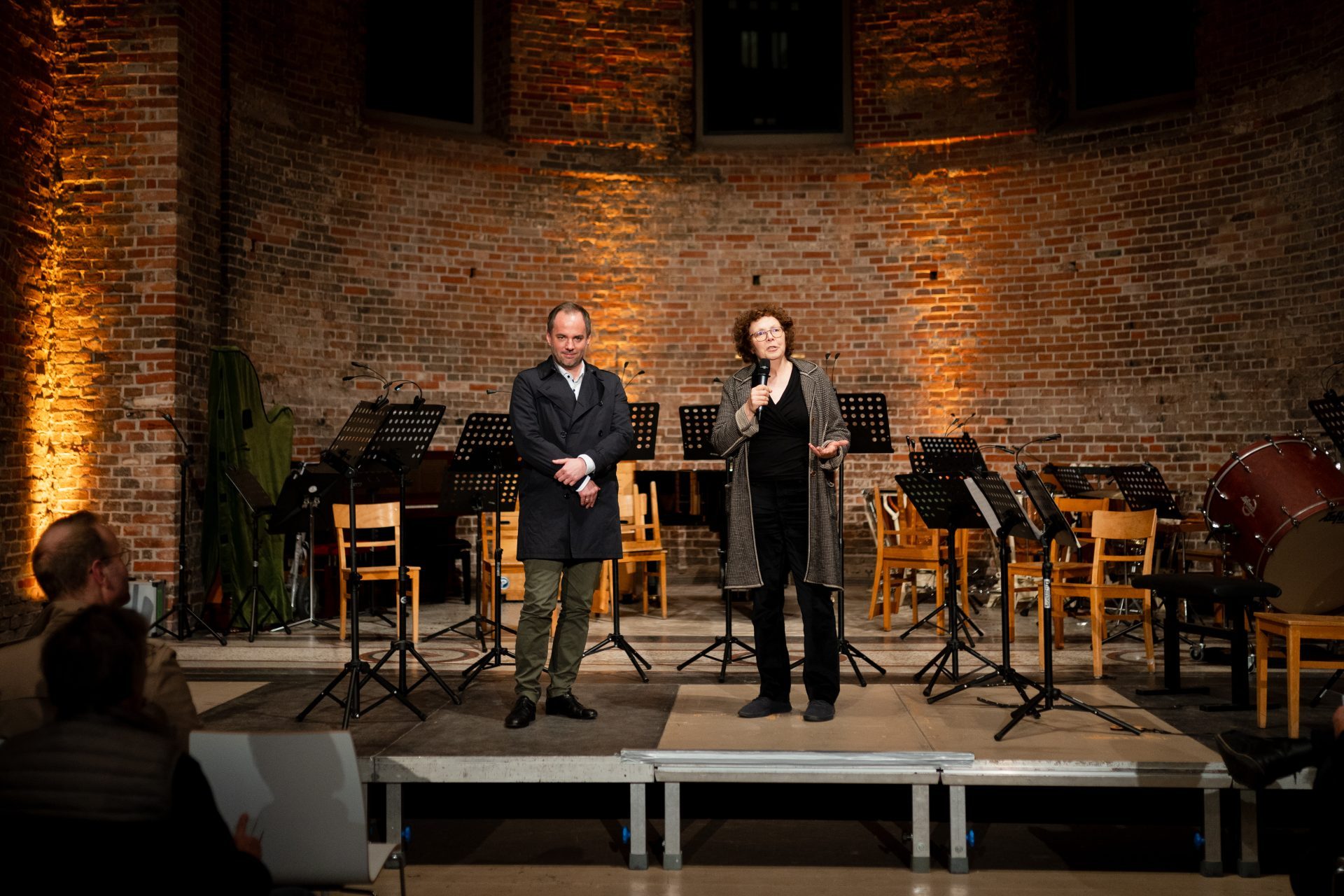 Zwei Personen stehen auf einer Bühne mit Musikinstrumenten und sprechen in einem gefliesten Raum mit warmem Licht.