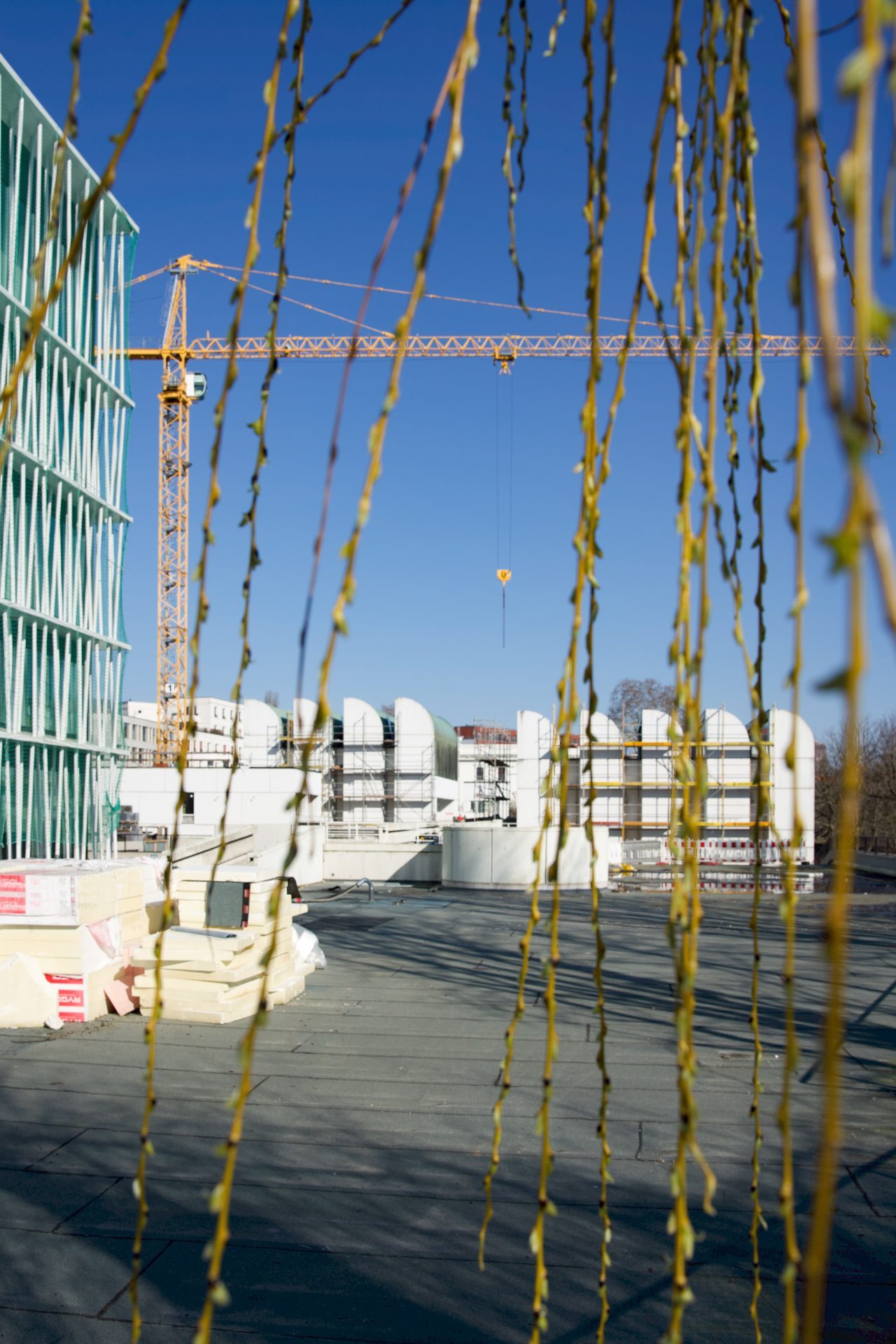 Foto des Baugeländes mit Turm, Kran und Baustellenmaterial, umgeben von grünen Zweigen und klarem blauen Himmel.