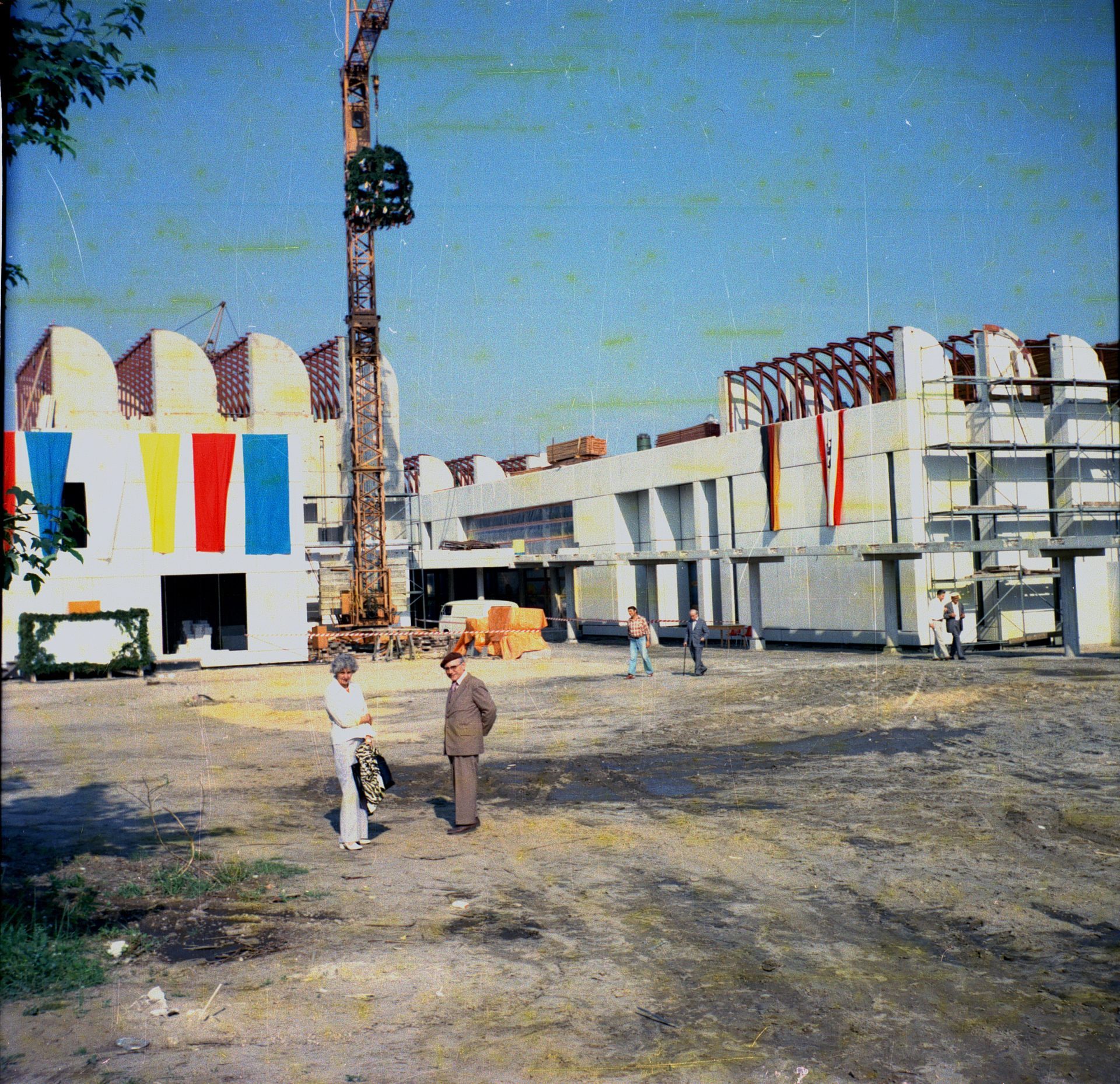 Foto des Bauplatzes mit Kran, davor sechs Besuchende, bunte Flaggen an Fassade in Gelb, Rot, Blau