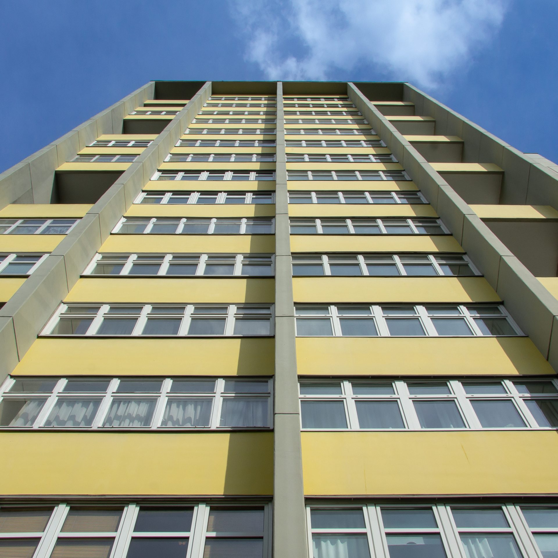 Ansicht eines hohen, gelben Wohnhauses mit zahlreichen Fenstern unter einem blauen Himmel.