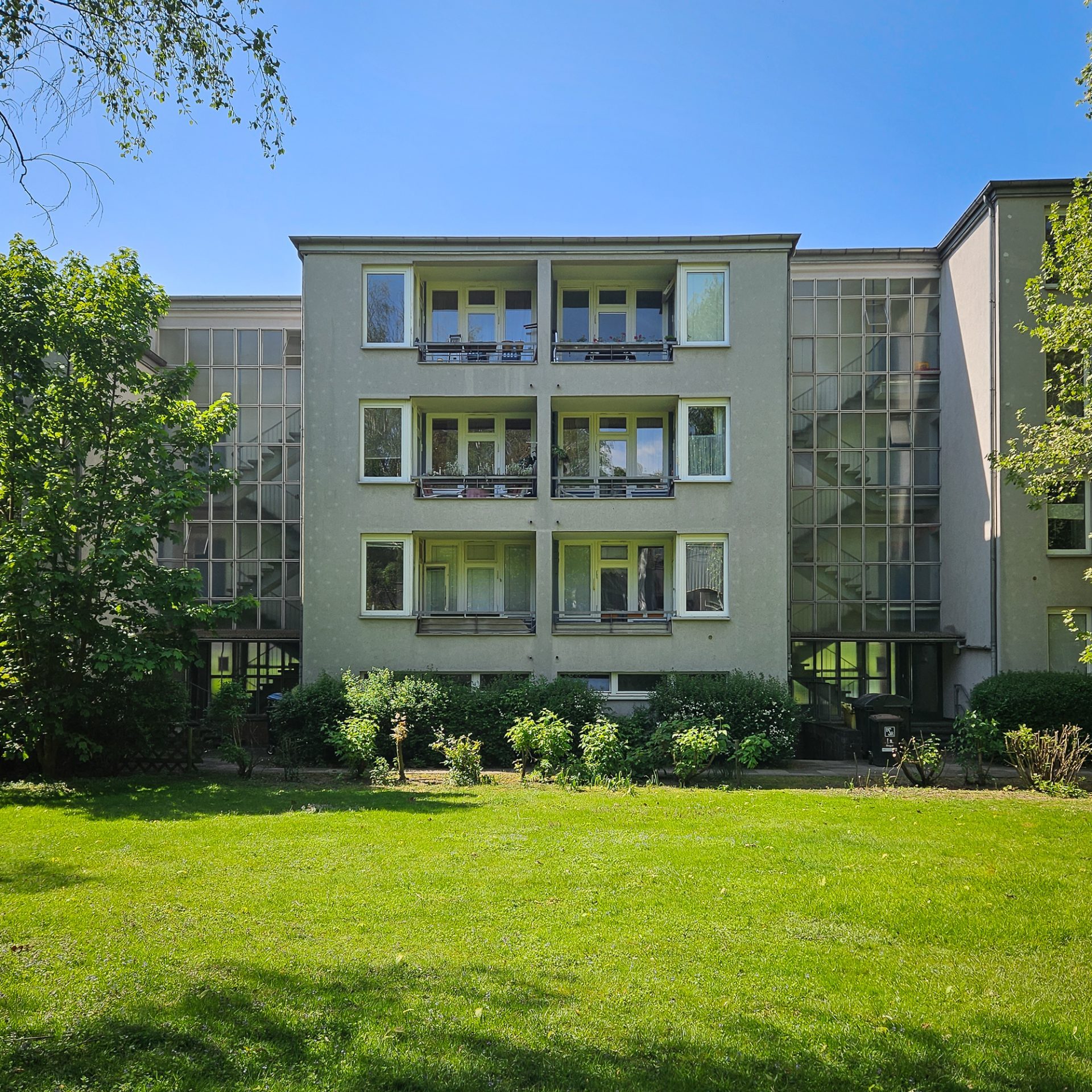 Moderne Wohngebäude mit Balkonen, umgeben von grünem Rasen und Bäumen unter einem strahlend blauen Himmel.