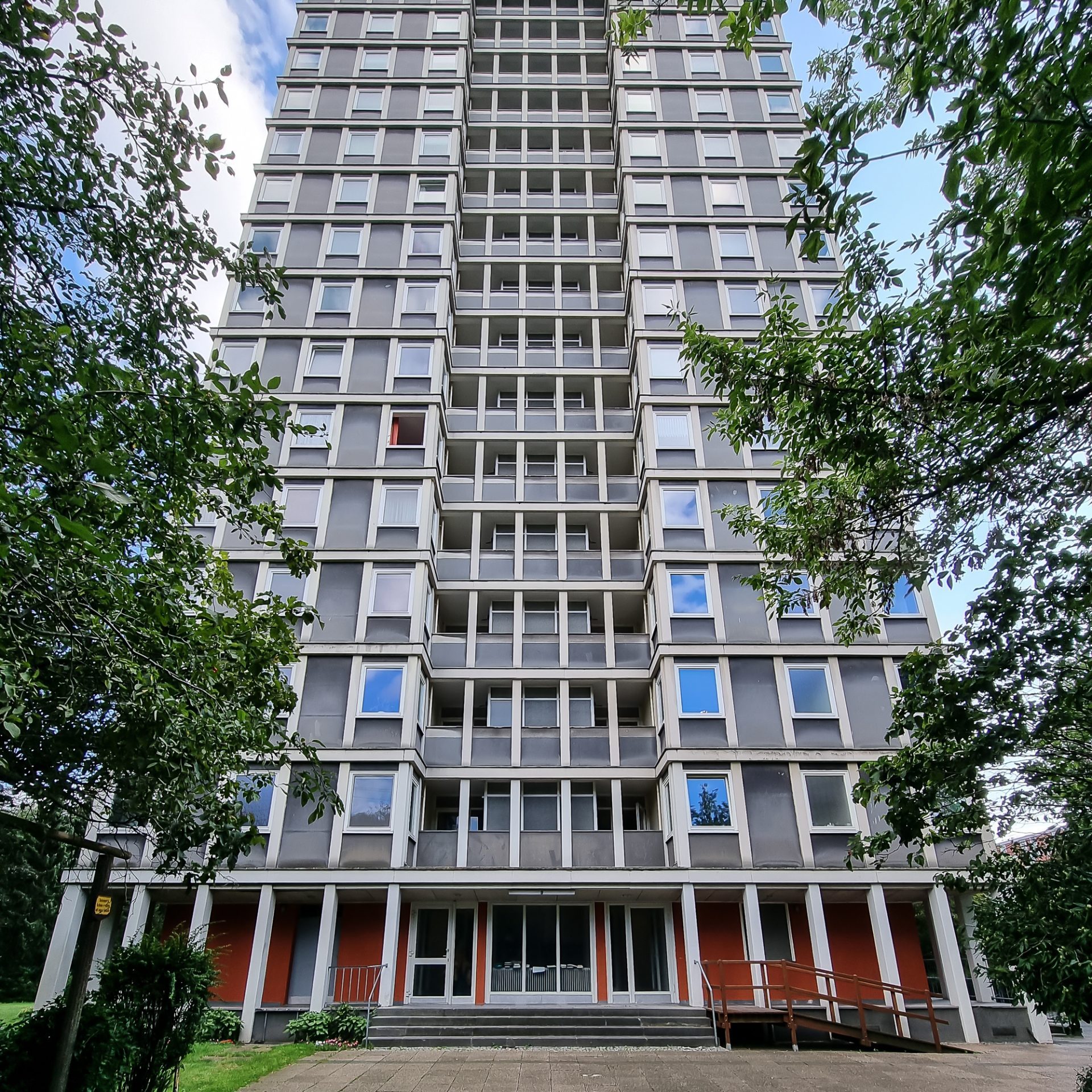 Hohes Wohngebäude mit grauer Fassade, umgeben von Bäumen, unter klarem blauen Himmel.