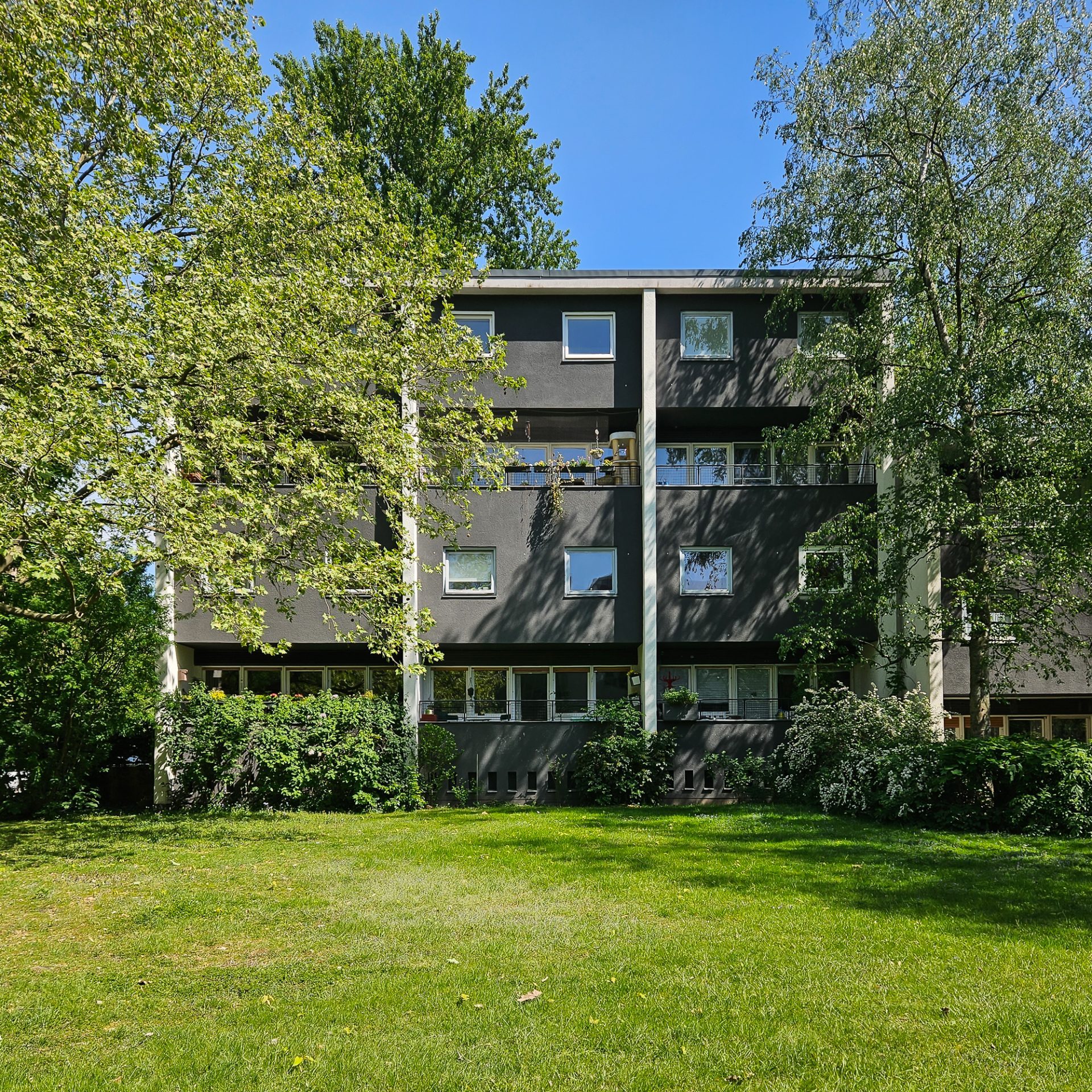 Moderner Wohnblock, umgeben von Bäumen und Grün, unter einem klaren blauen Himmel.