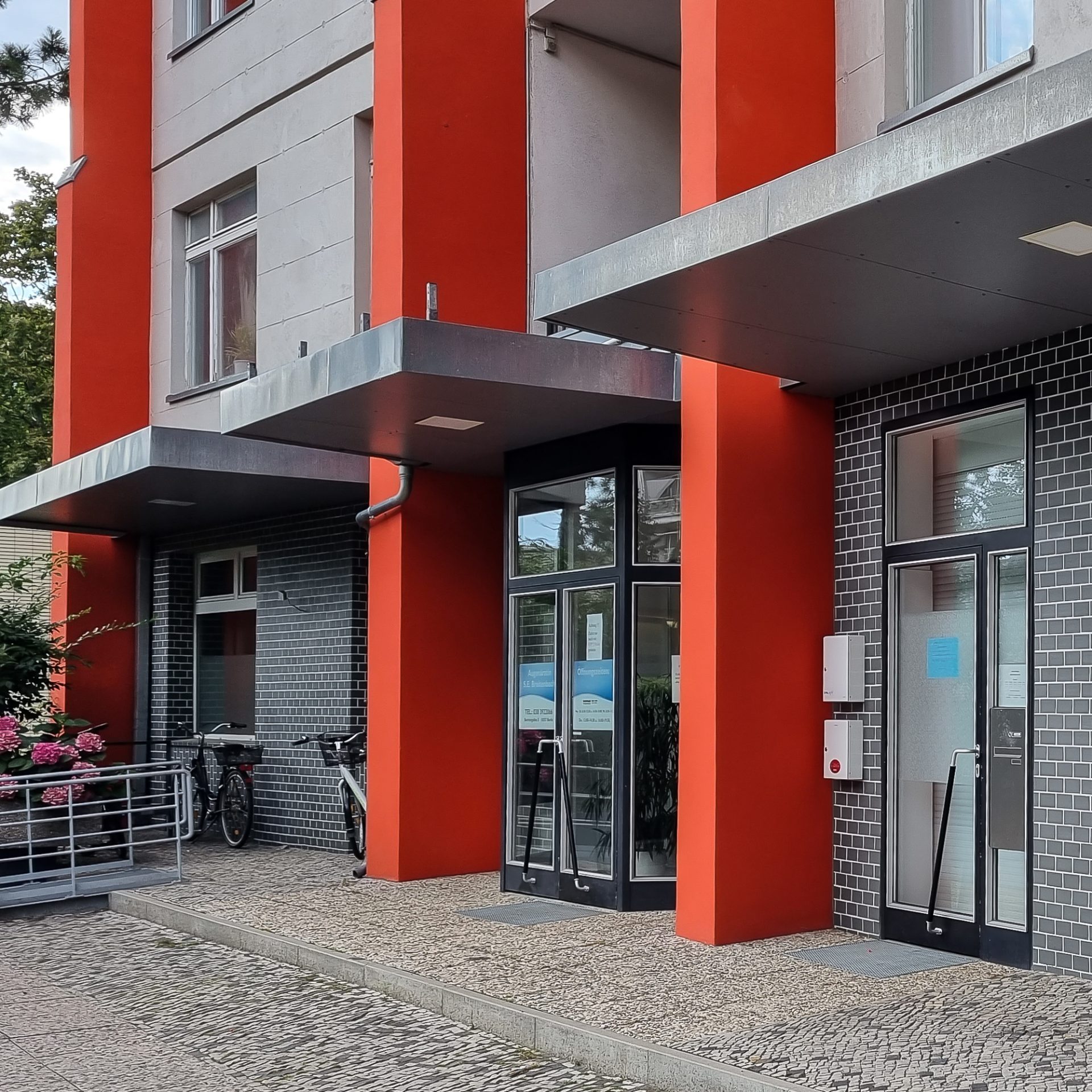Eingang zu einem modernen Gebäude mit rotem Anstrich und Glasportalen, umgeben von Pflanzen und Fahrrädern.