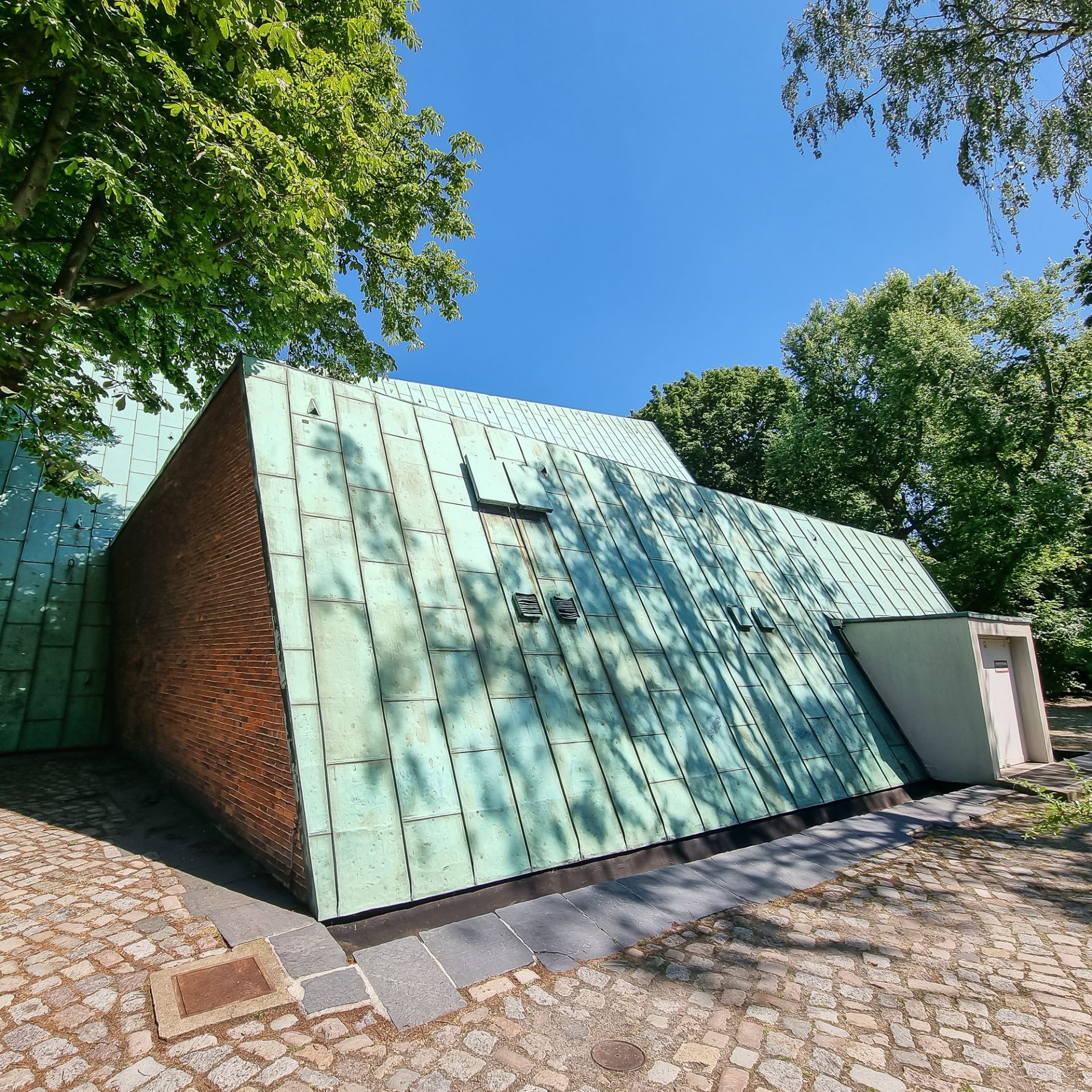 Moderne Architektur mit grünem Metalldach und Ziegelwand, umgeben von Bäumen unter blauem Himmel.
