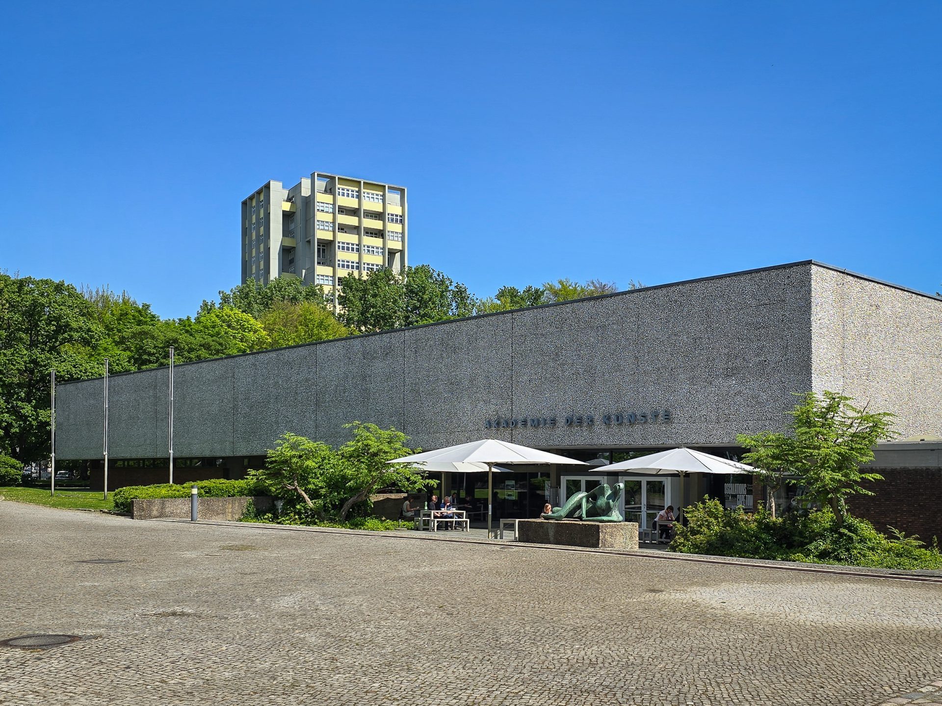 Modernes Gebäude mit flacher Fassade, begrünten Flächen und Sonnenschirmen, im Hintergrund ein höheres Gebäude unter blauem Himmel.