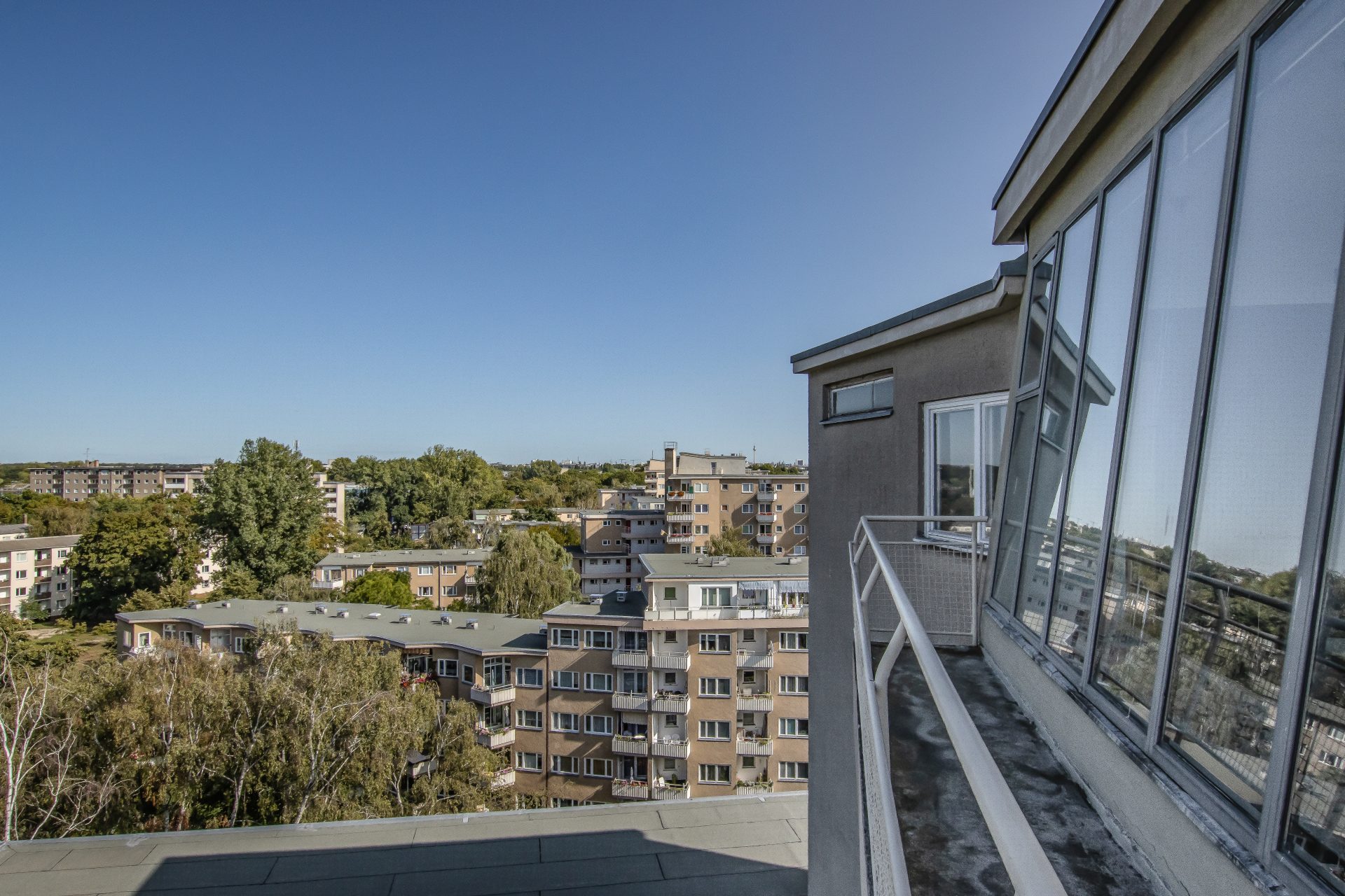 Fotografie, Blick von einem Balkon auf ein Stadtviertel mit Wohngebäuden und klarem, blauem Himmel.