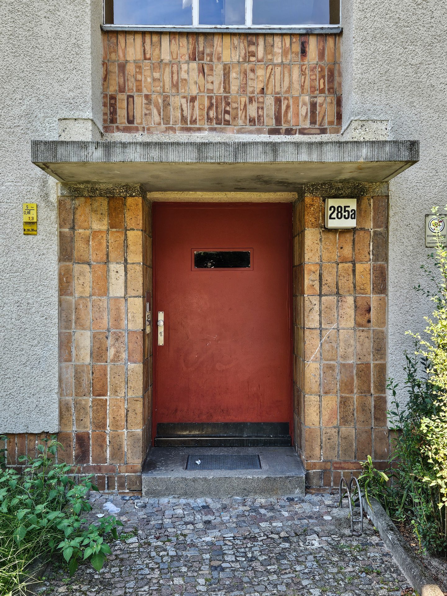 Rote Eingangstür mit schmalem Fenster, umgeben von hellen Ziegeln und Pflanzen. Hausnummer 285a sichtbar.