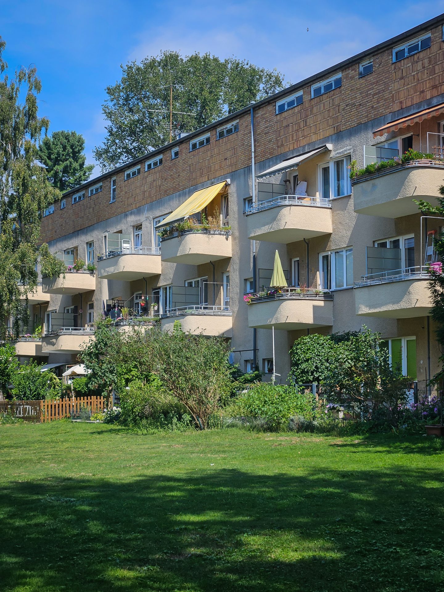 Mehrstöckiges Wohngebäude mit Balkonen, umgeben von grünem Rasen und Bäumen an einem sonnigen Tag.