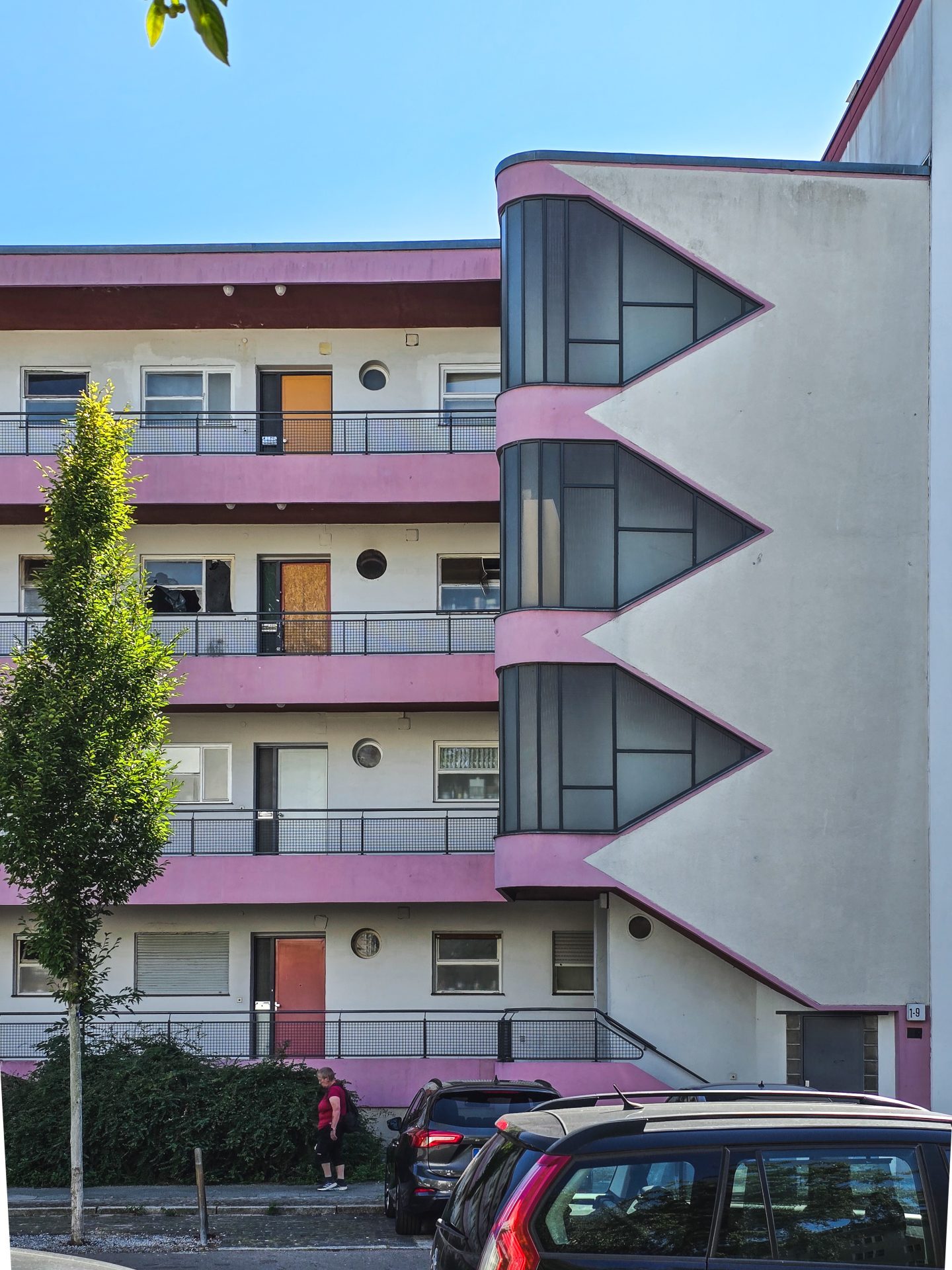 Modernes Wohngebäude mit pinken Akzenten, Glasfront und begrünten Parkplätzen unter blauem Himmel.