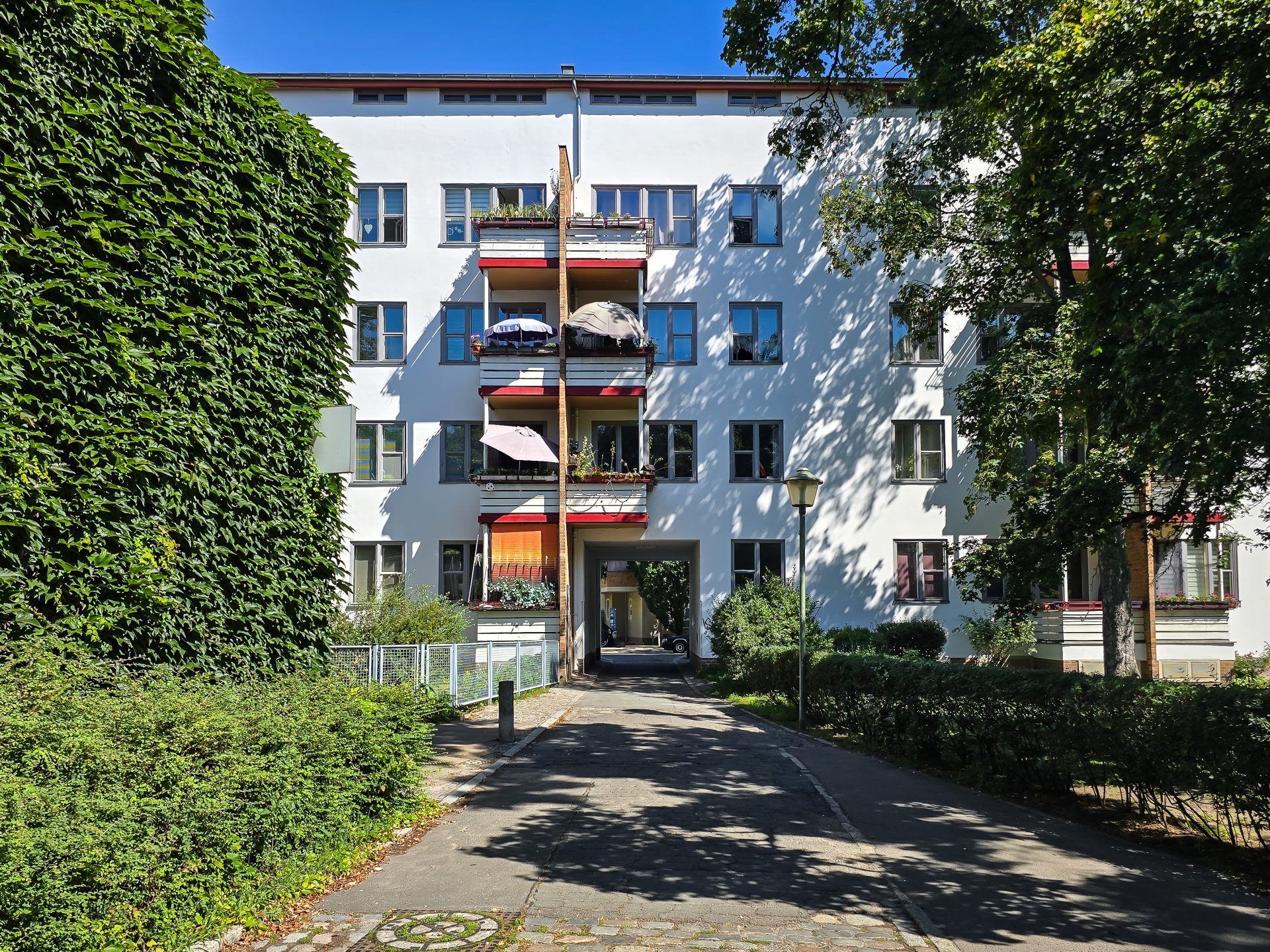 Weißes Wohnhaus mit Balkonen, umgeben von Bäumen und Sträuchern, bei klarem, sonnigem Wetter.