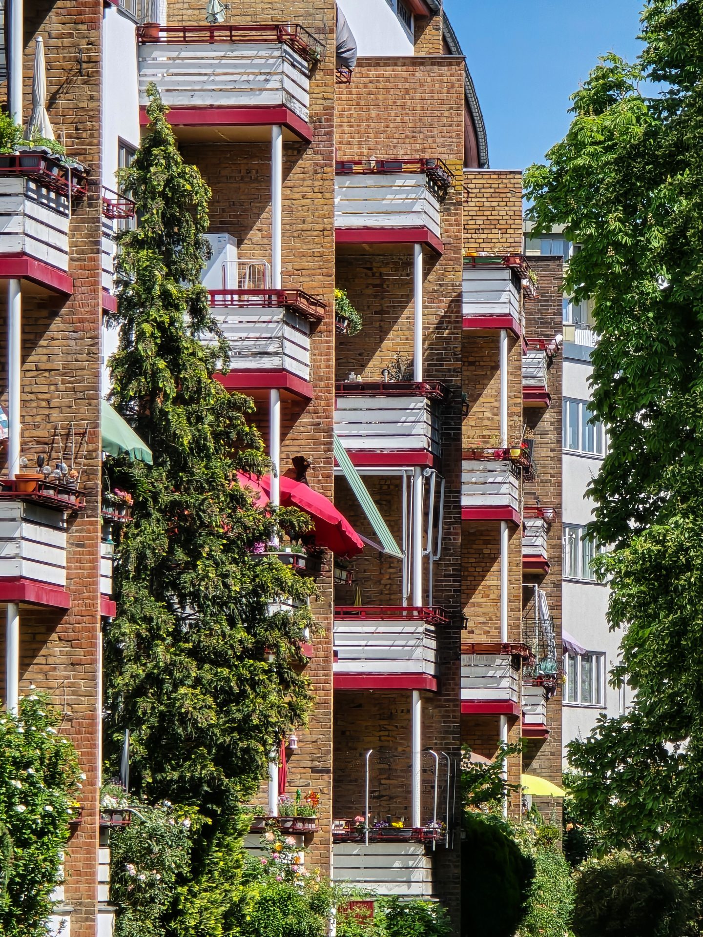 Bunte Balkone an einem Ziegelgebäude, umgeben von grünen Bäumen und strahlend blauem Himmel.