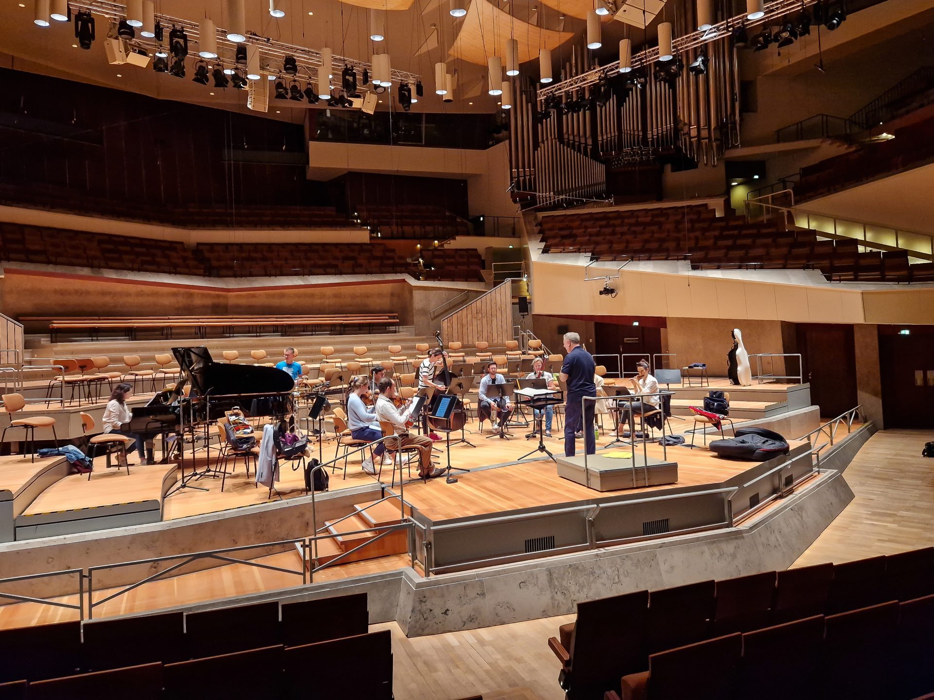 Orchesterproben auf einer Bühne in einem modernen Saal, mit Klavier und mehreren Musikern.