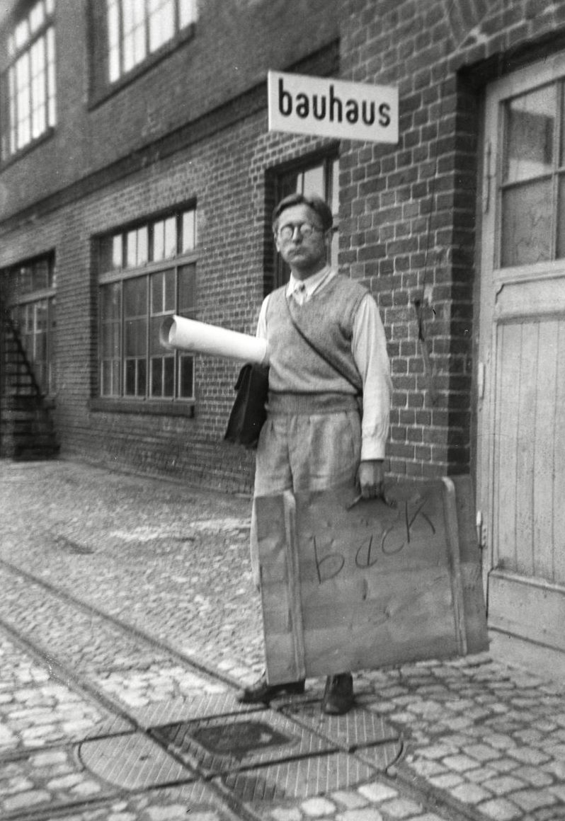 Schwarz-Weiß-Fotografie einer Person mit kurzen Haaren vor dem Bauhaus-Gebäude mit Schild, Entwurf unter dem Arm