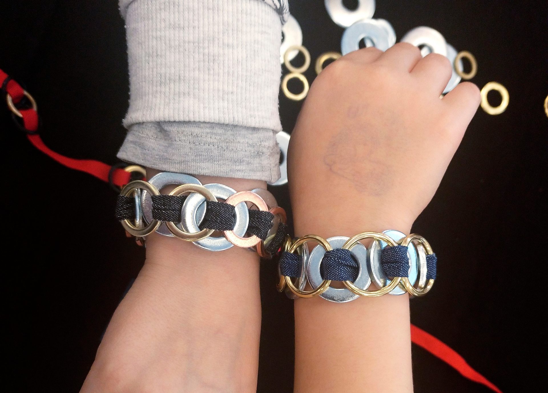 Zwei Handgelenke mit bunten, selbstgemachten Armbändern aus Metallringen und Stoff auf schwarzem Hintergrund.