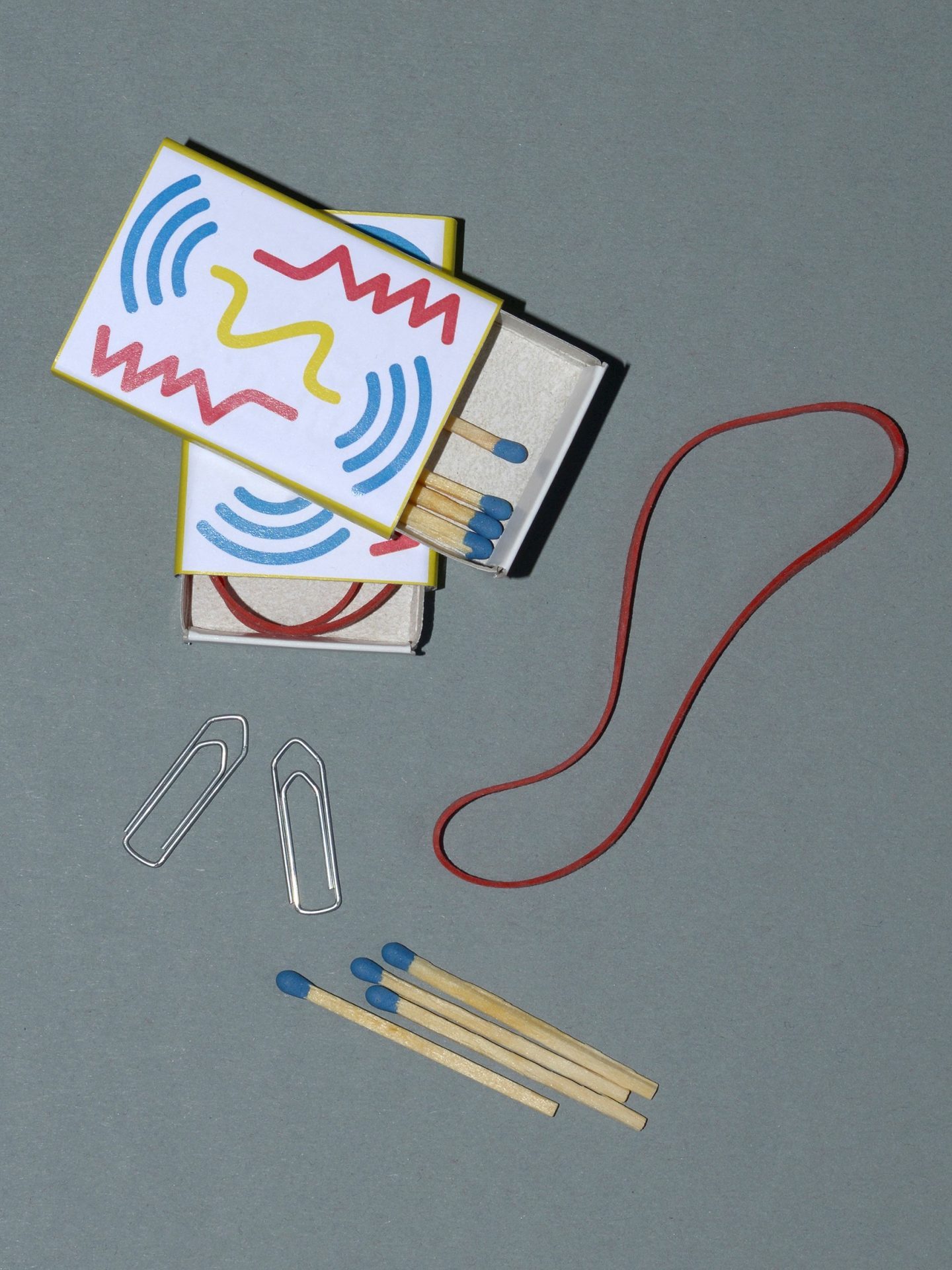 Streichholzschachtel mit buntem Design, Streichhölzer, Büroklammern und ein Gummiband auf grauem Hintergrund.