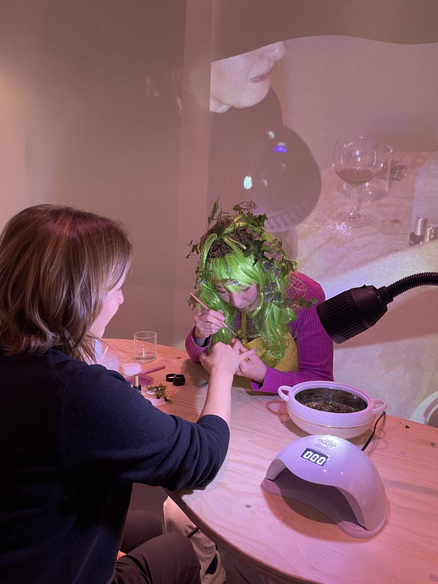A woman with green hair and leaf decor gets a manicure while another woman works in the background.
