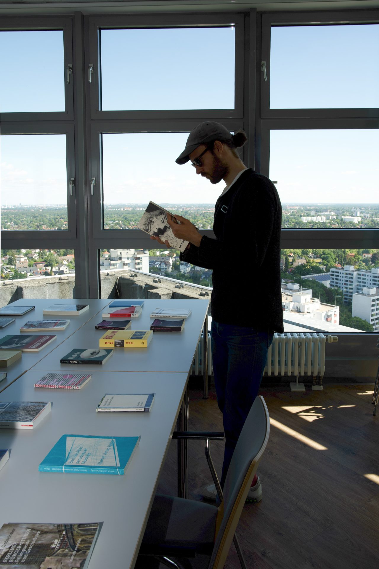 Mann steht an einem Tisch mit Büchern, liest ein Buch, im Hintergrund Blick auf die Stadt.