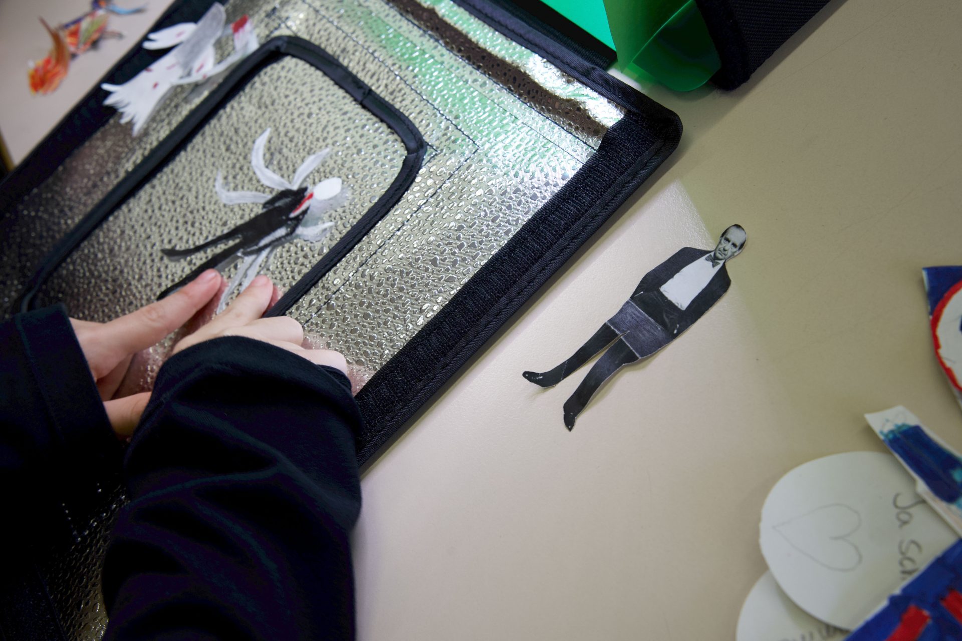 Hände eines Kindes spielen mit Papierfiguren auf einer reflektierenden Oberfläche, umgeben von bunten Bastelmaterialien.