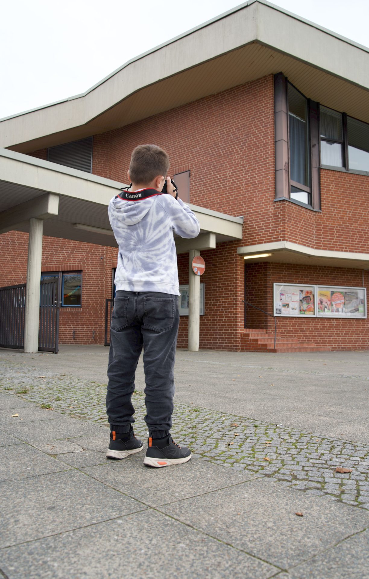 Ein Junge mit einer Kamera fotografiert an einem grauen Tag ein rotes Ziegelsteingebäude.