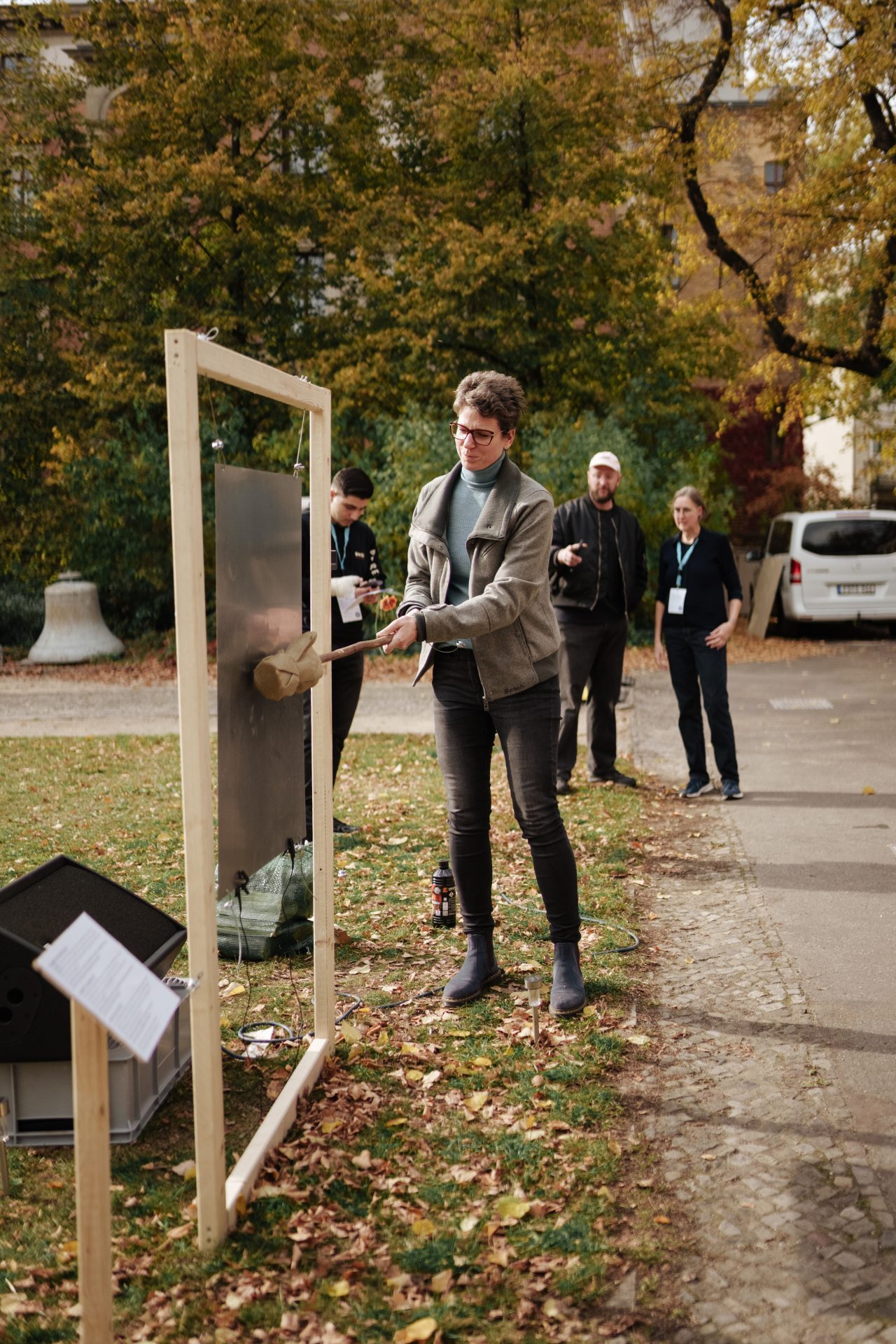 Eine Person schlägt mit einem Werkzeug auf ein Metallobjekt, während Zuschauer im Hintergrund stehen. Herbstliche Szenerie.