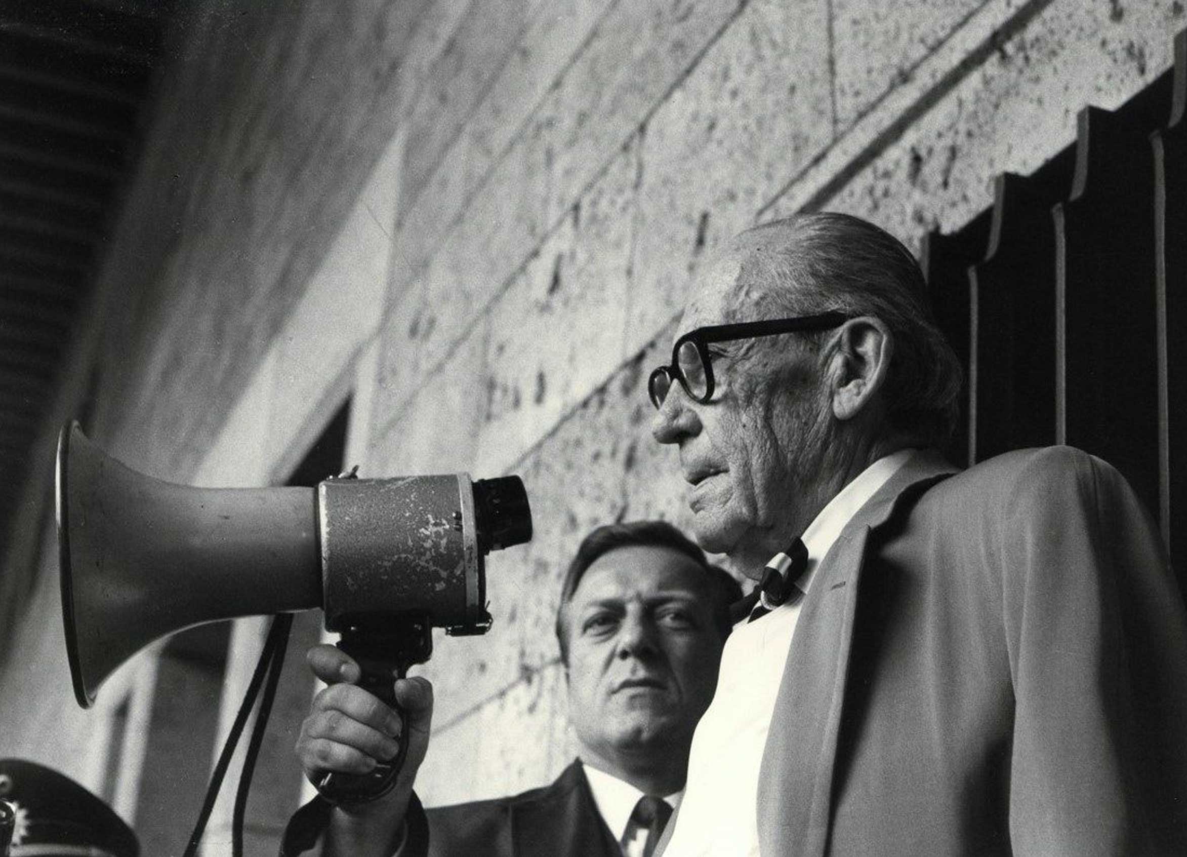 Two men speaking, one holding a megaphone, in a black and white setting.