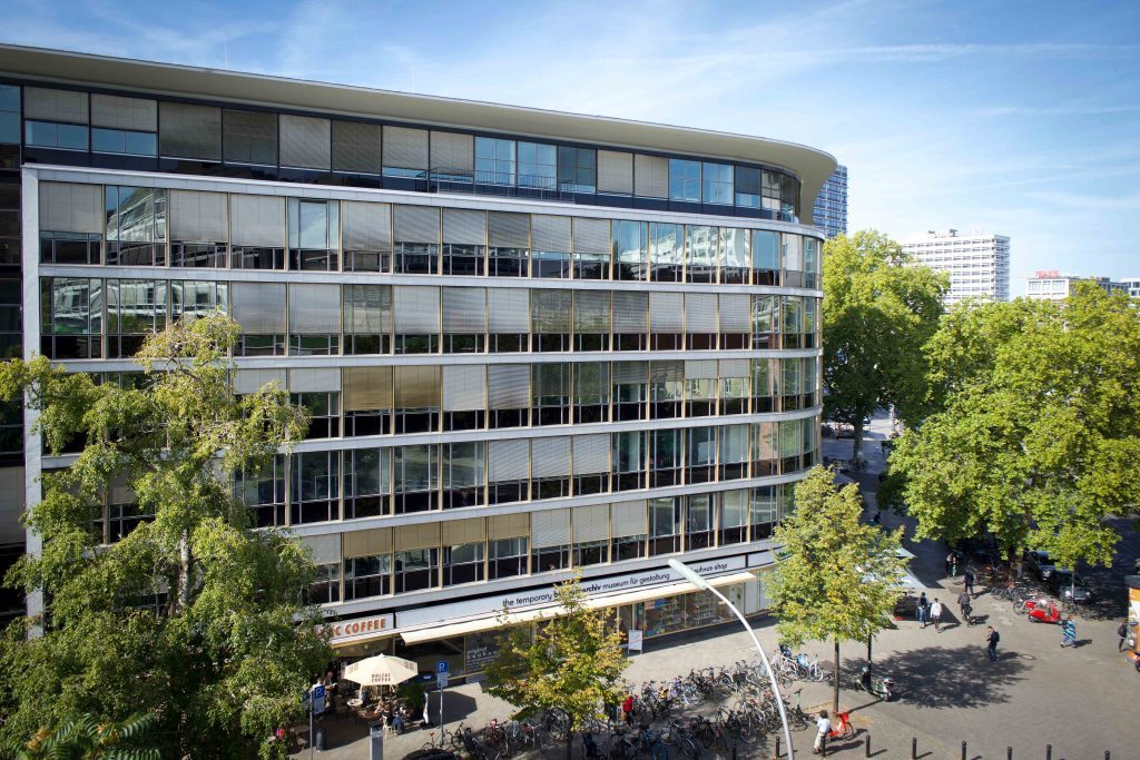 Modern building with large windows, trees, and a café on the ground floor, lively street with bicycles and pedestrians.
