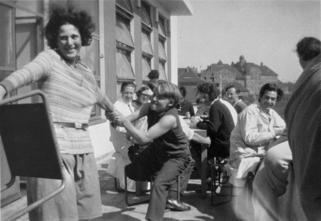 Two women dance joyfully outdoors while others enjoy a social gathering at tables in the background.
