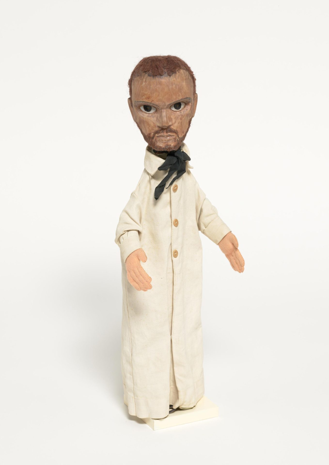 Holzfigur mit braunem Haar, Bart, Brille und weißem Gewand, die auf einem Sockel steht.