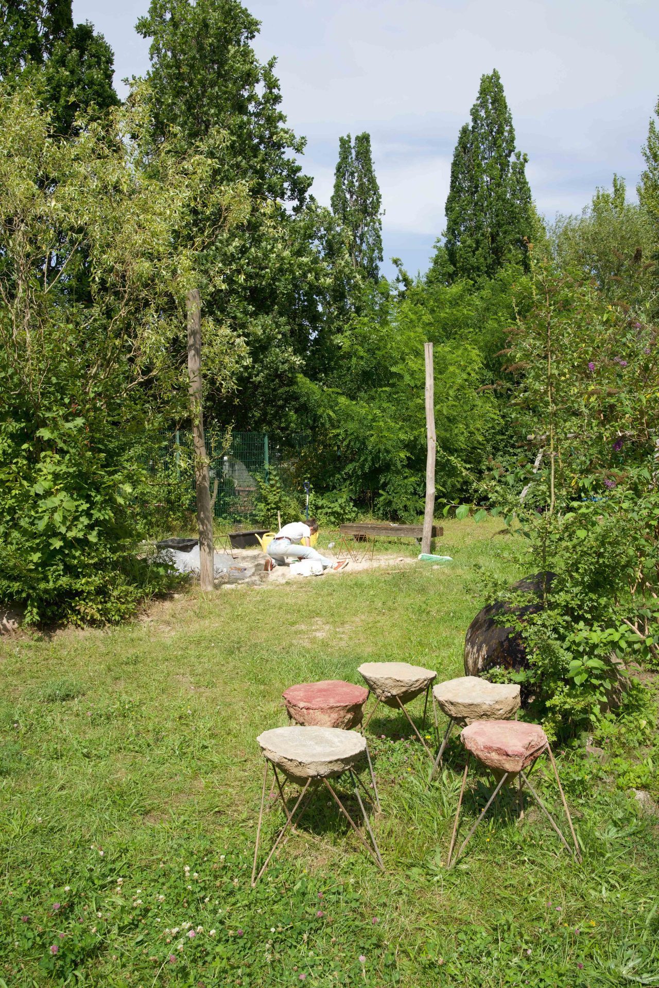 Ein grüner Garten mit vier Stein-Sitz- und Stehgelegenheiten sowie einem Arbeitsbereich im Hintergrund.