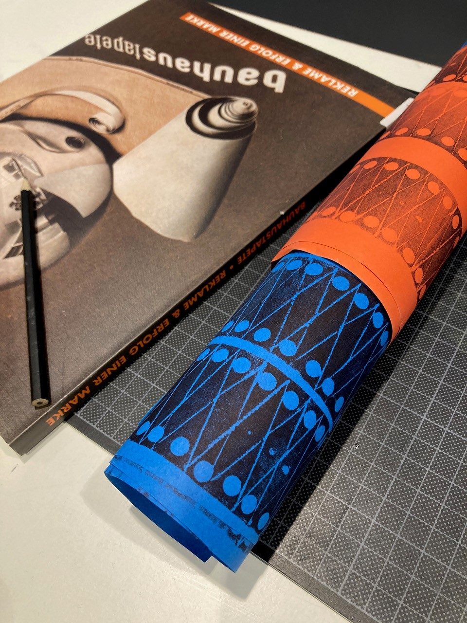 Bunte Papierrolle in Blau und Orange, daneben ein Bauhaus-Buch und ein Stift auf einem grafischen Hintergrund.
