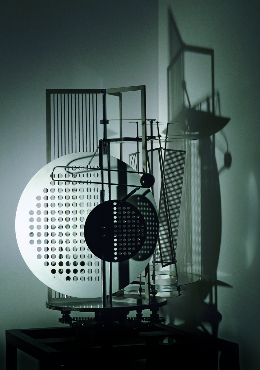 Abstrakte Metallskulptur mit durchbrochenen Flächen, die komplexe Schatten an die Wand wirft.