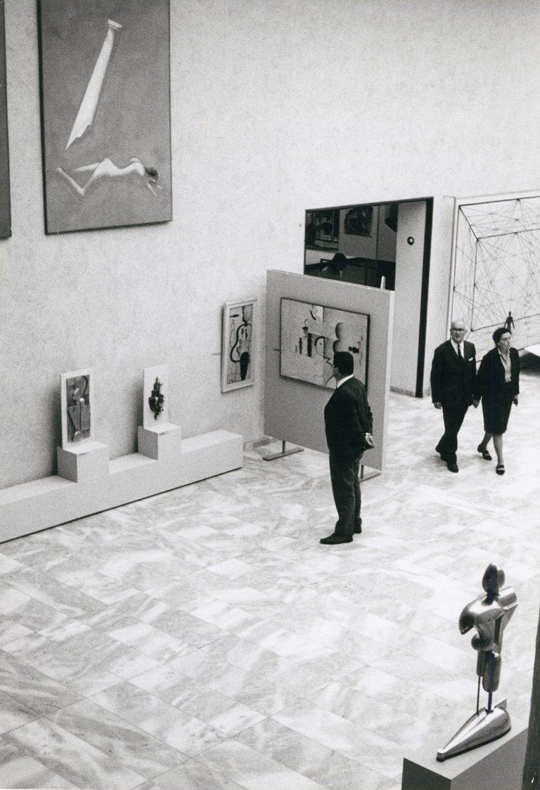 Schwarz-Weiß-Fotografie, drei Besuchende im einem hohen und marmorierten Ausstellungsraum betrachten Kunstwerke und Skulpturen.