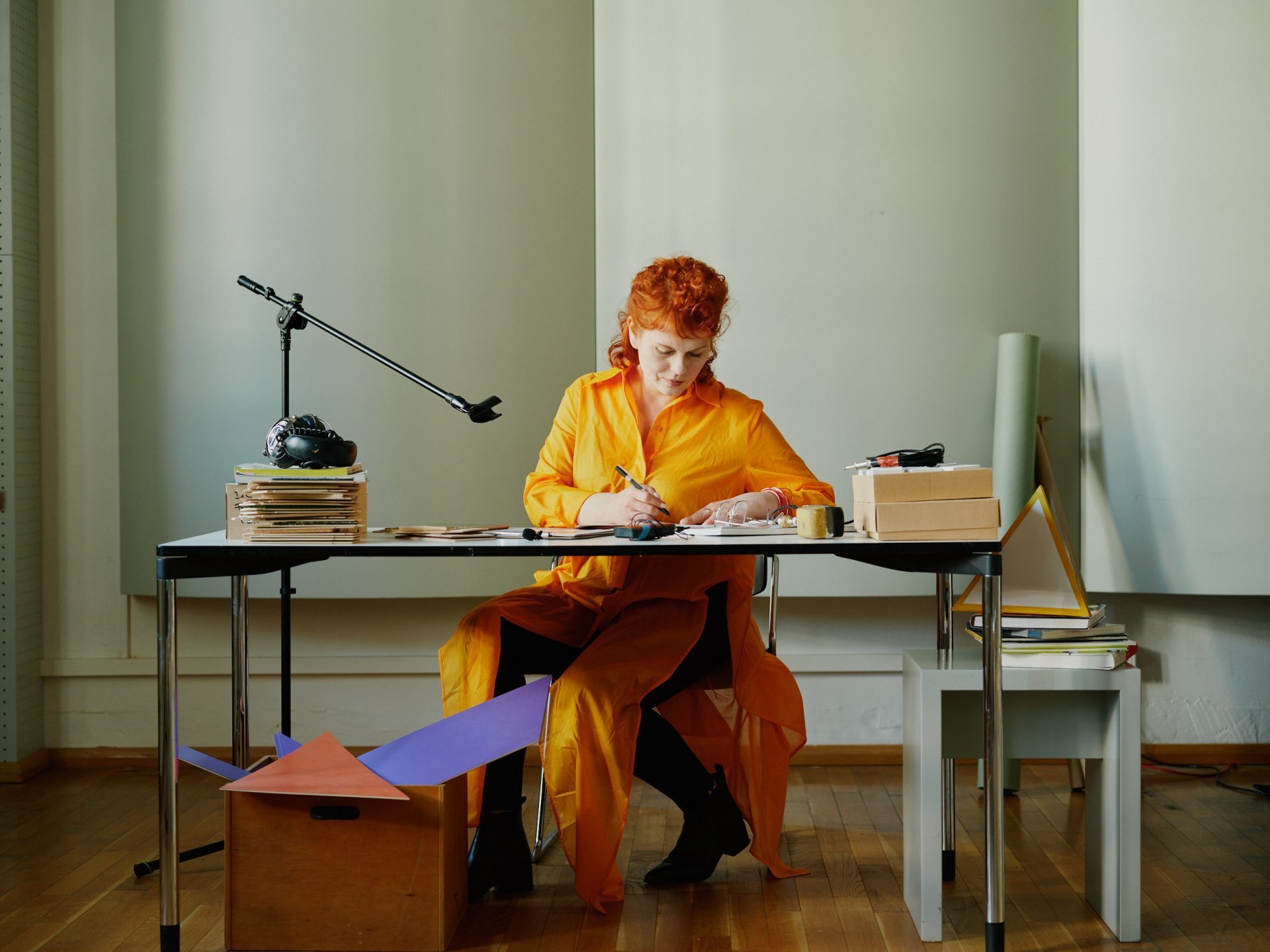 Eine Person mit roten Haaren in einem orangefarbenen Outfit arbeitet an einem Tisch mit diversen Materialien.