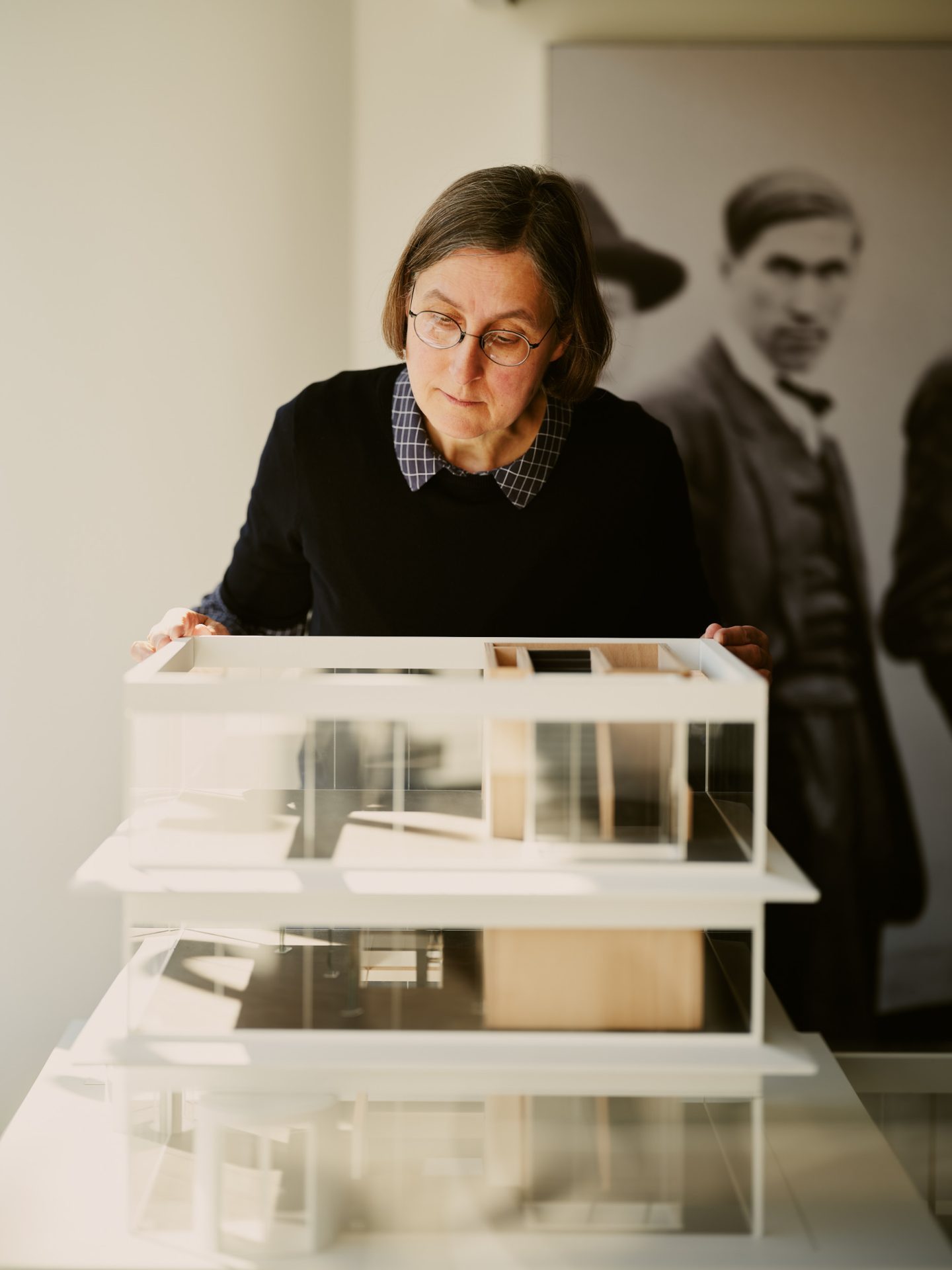 Eine Frau begutachtet ein Architekturmodell in einem hellen Raum, im Hintergrund sind historische Fotos sichtbar.