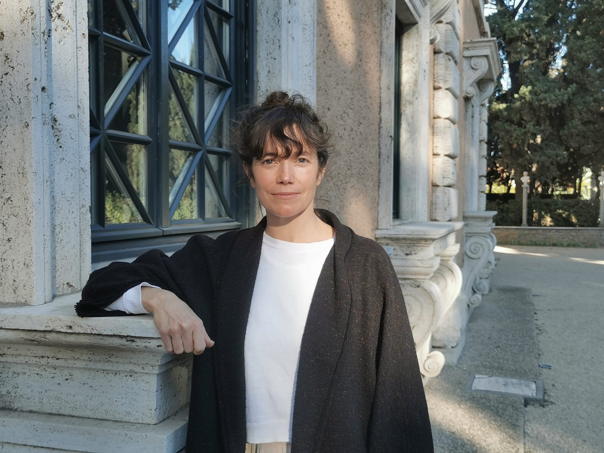 Frau mit lockigem Haar an einer alten Wand, umgeben von Grün und historischen Architekturdetails.