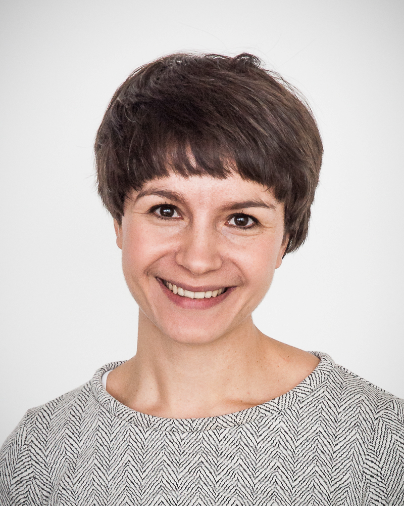 Lächelnde Frau mit kurzen Haaren in einem grauen Oberteil vor hellem Hintergrund.
