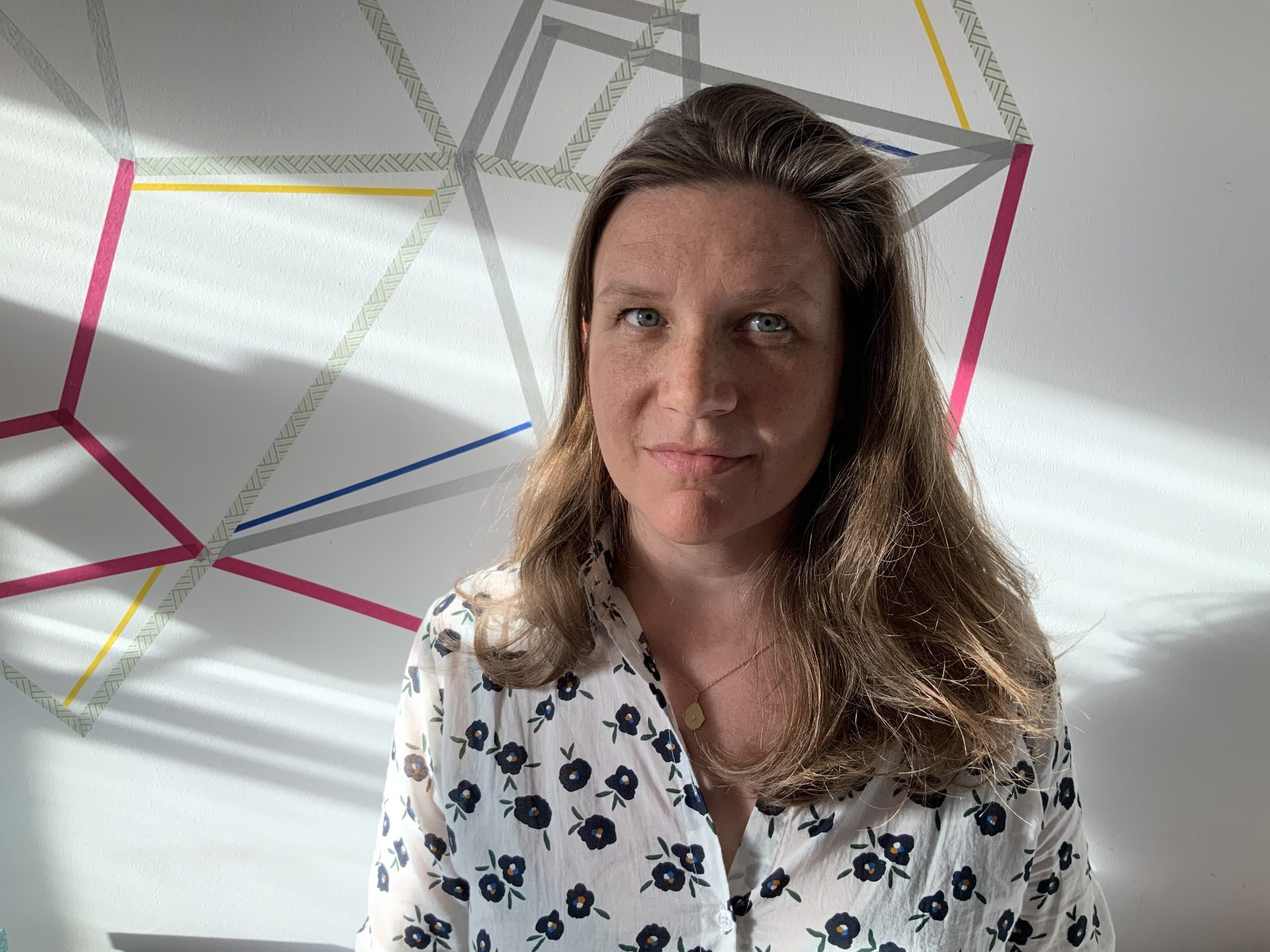 Frau mit langen Haaren in gemustertem Hemd vor einer geometrischen Wandgestaltung mit Lichtspielen.