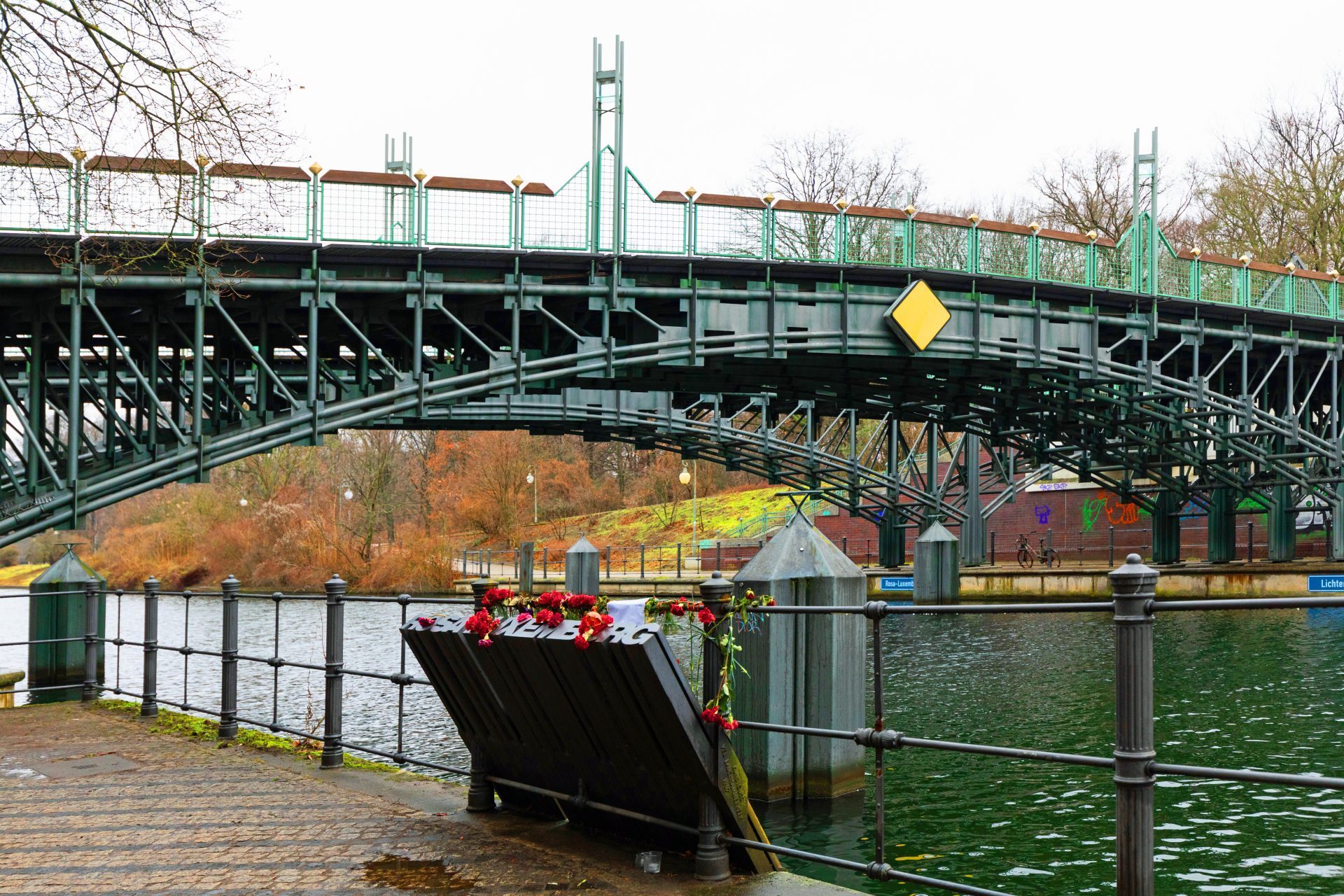 Eisenbahnbrücke über Wasser, umgeben von einem Blumenkranz am Ufer und herbstlichem Laub im Hintergrund.